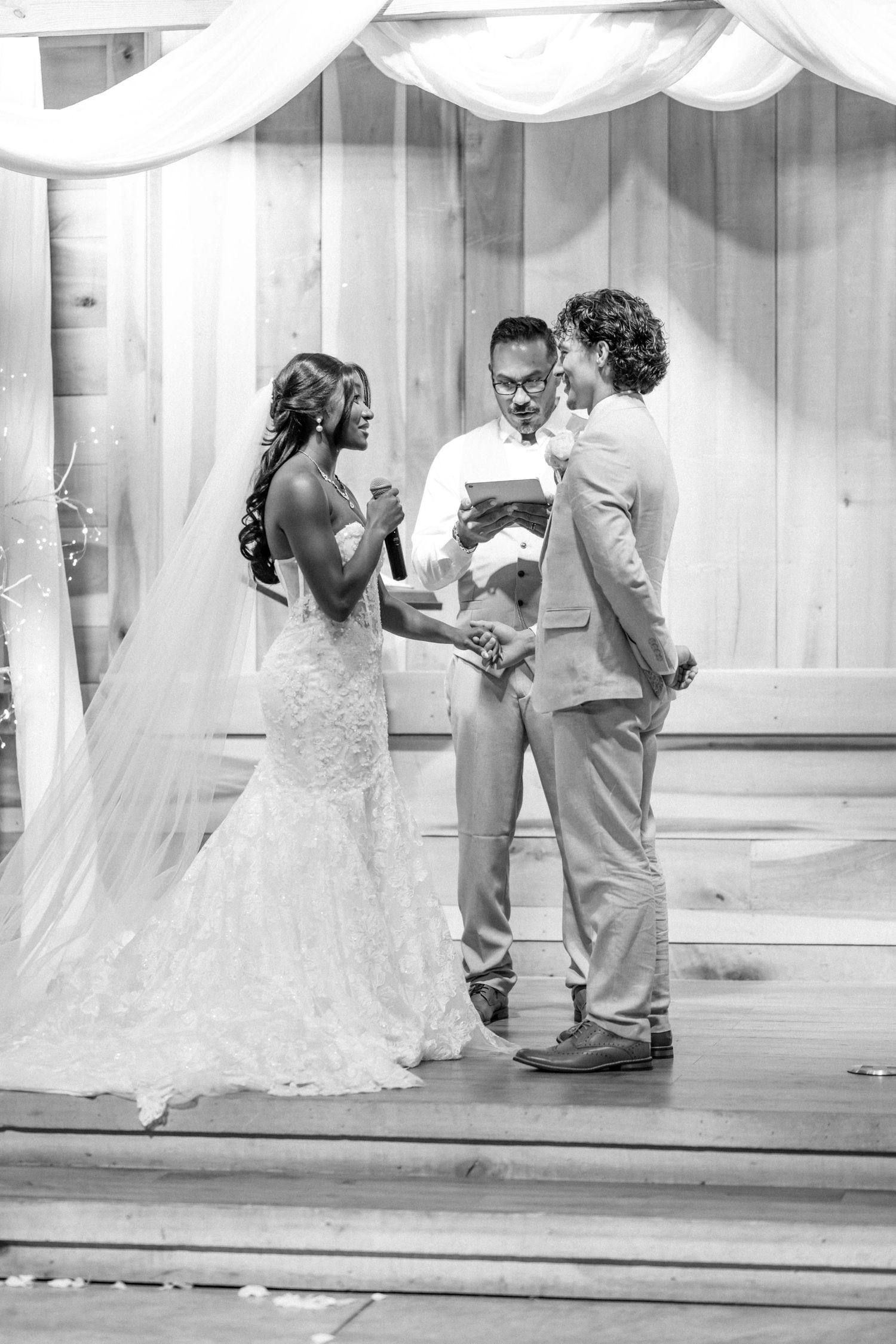 Black and white wedding ceremony photo of bride in lace gown exchanging vows with groom as officiant reads from booklet.