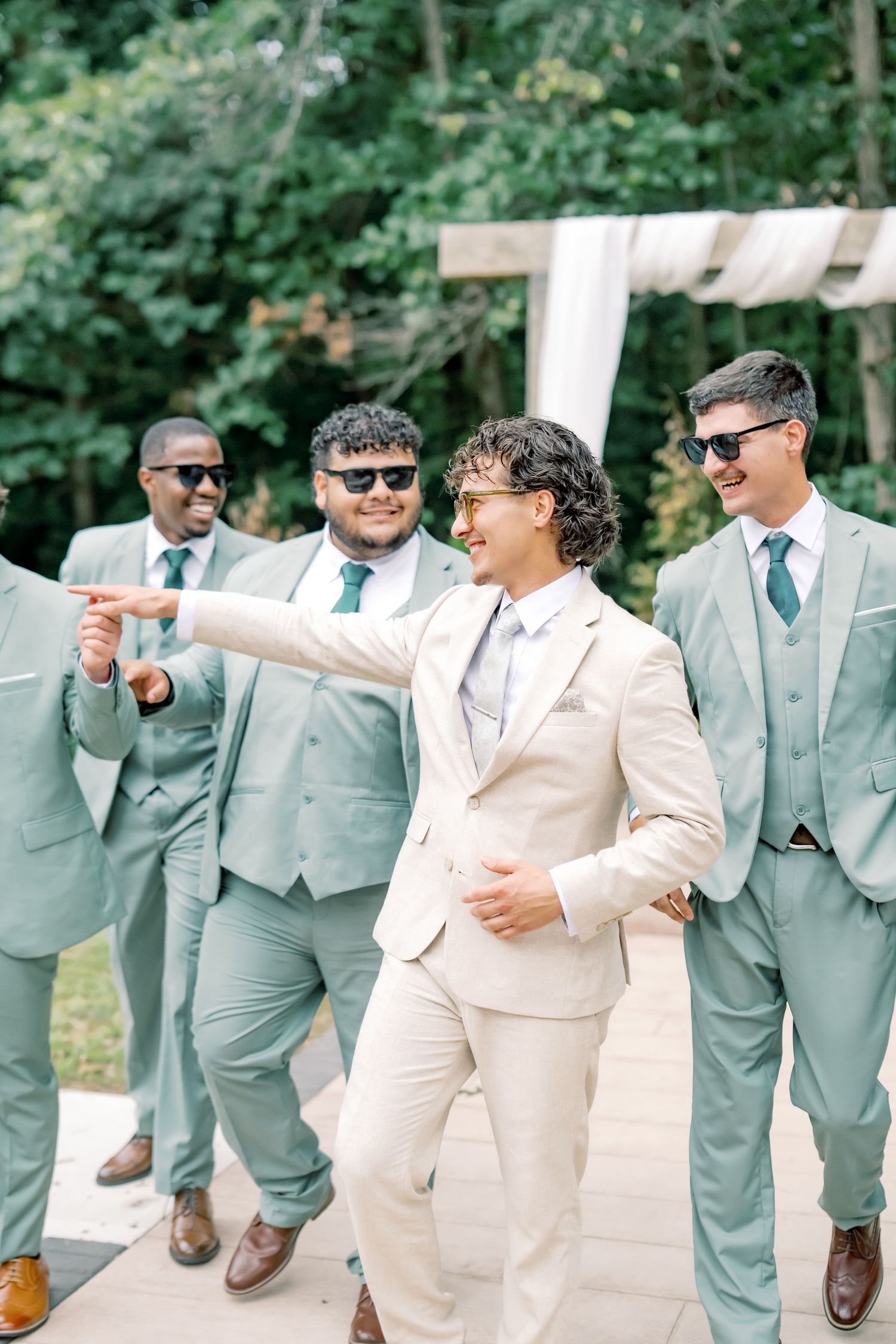 Groom in beige suit laughs with groomsmen in sage suits and sunglasses at outdoor wedding.