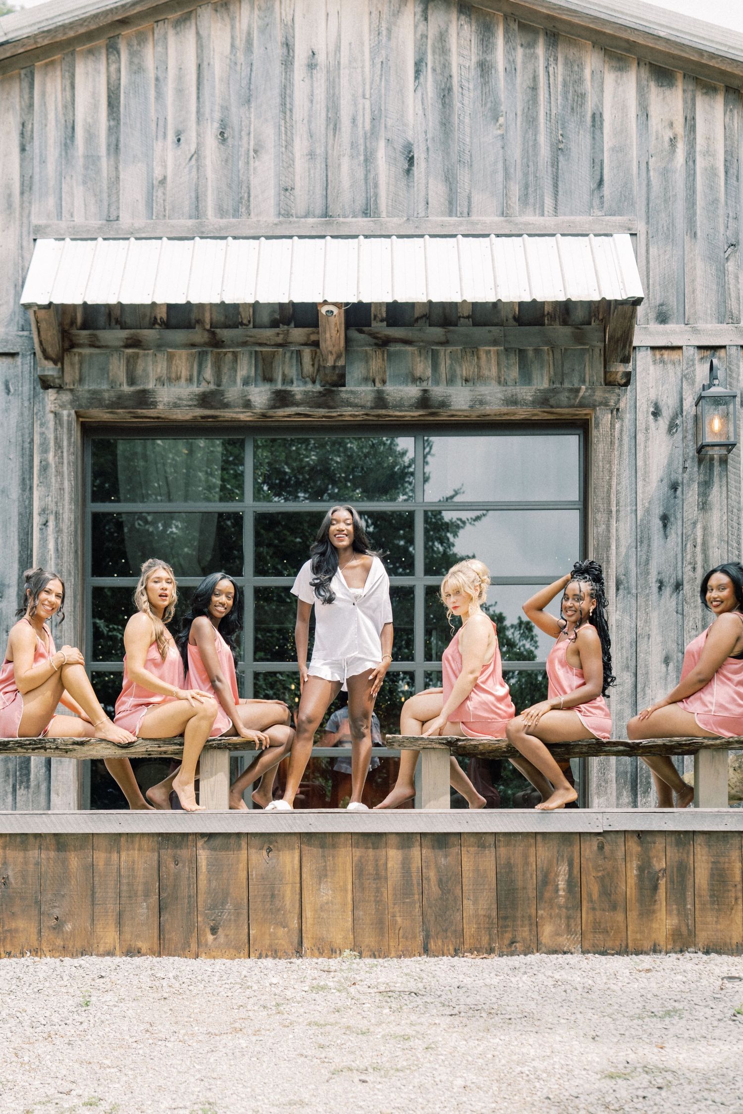 Bride in white robe stands with bridesmaids in pink robes on rustic barn porch for wedding day getting ready photos.