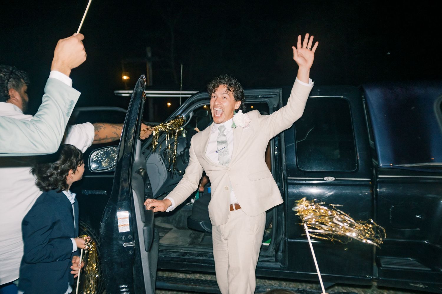 Groom in cream suit waves joyfully while exiting black limousine during festive wedding night celebration.