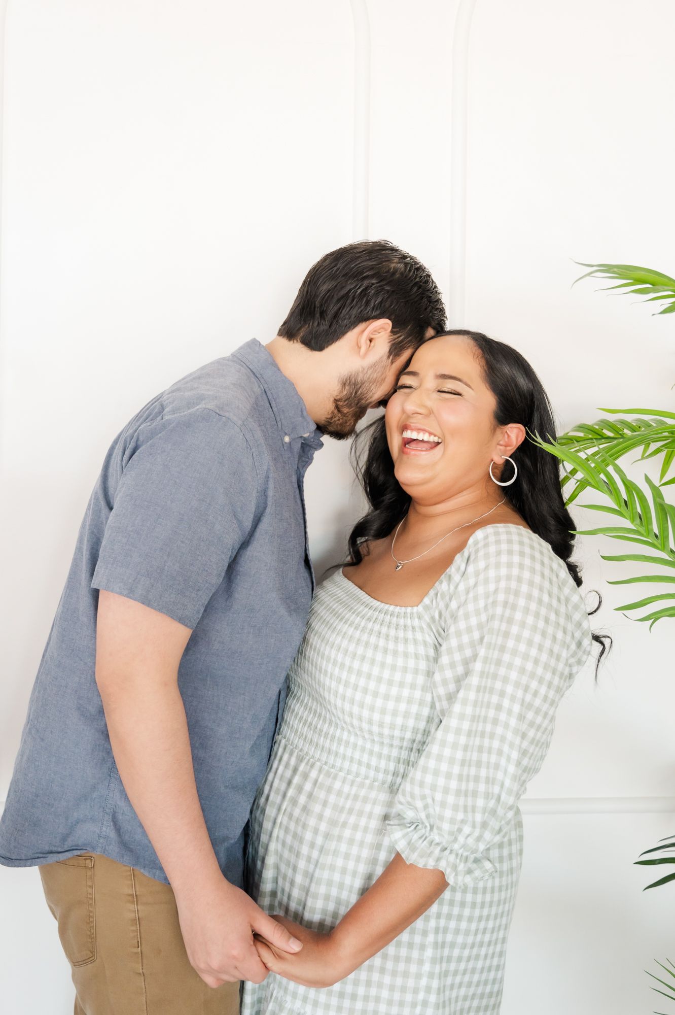 A couple shares an intimate moment and laughter against a white wall with tropical plants.