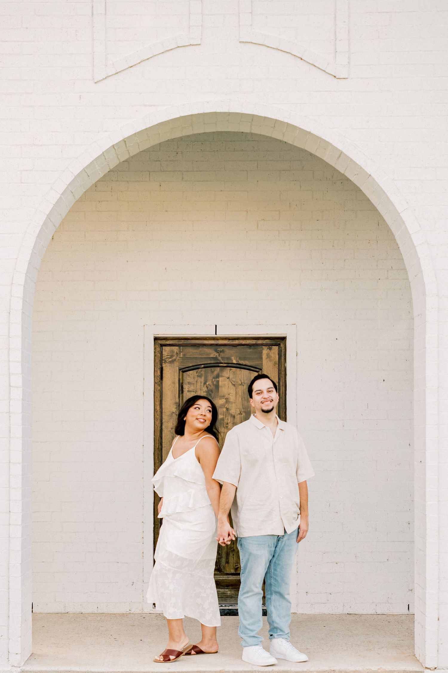 A series of white arched doorways frame a couple posing in casual attire against a minimalist architectural backdrop.