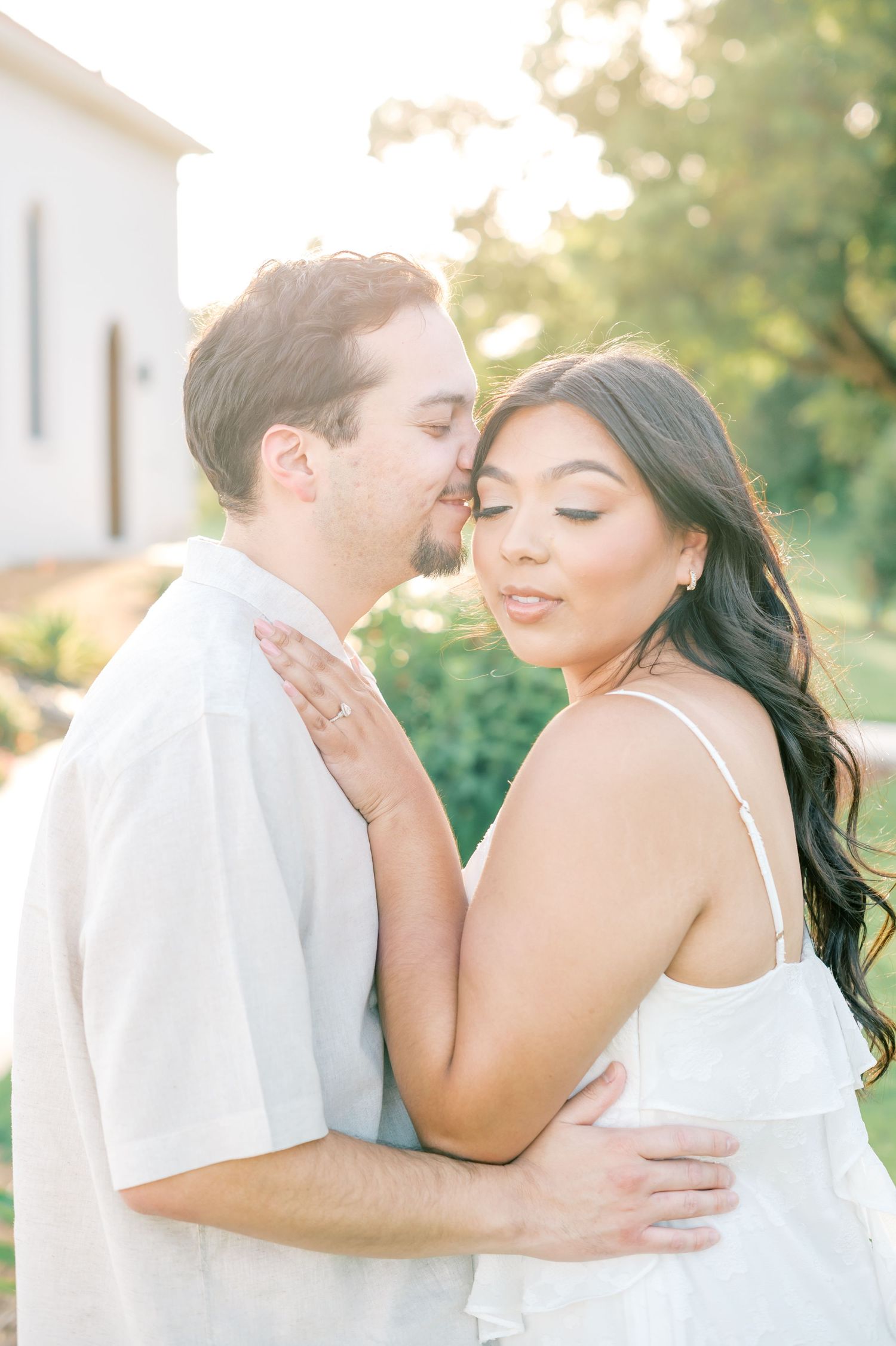 A romantic couple embraces outdoors in soft sunlight wearing white outfits during an engagement photo session.
