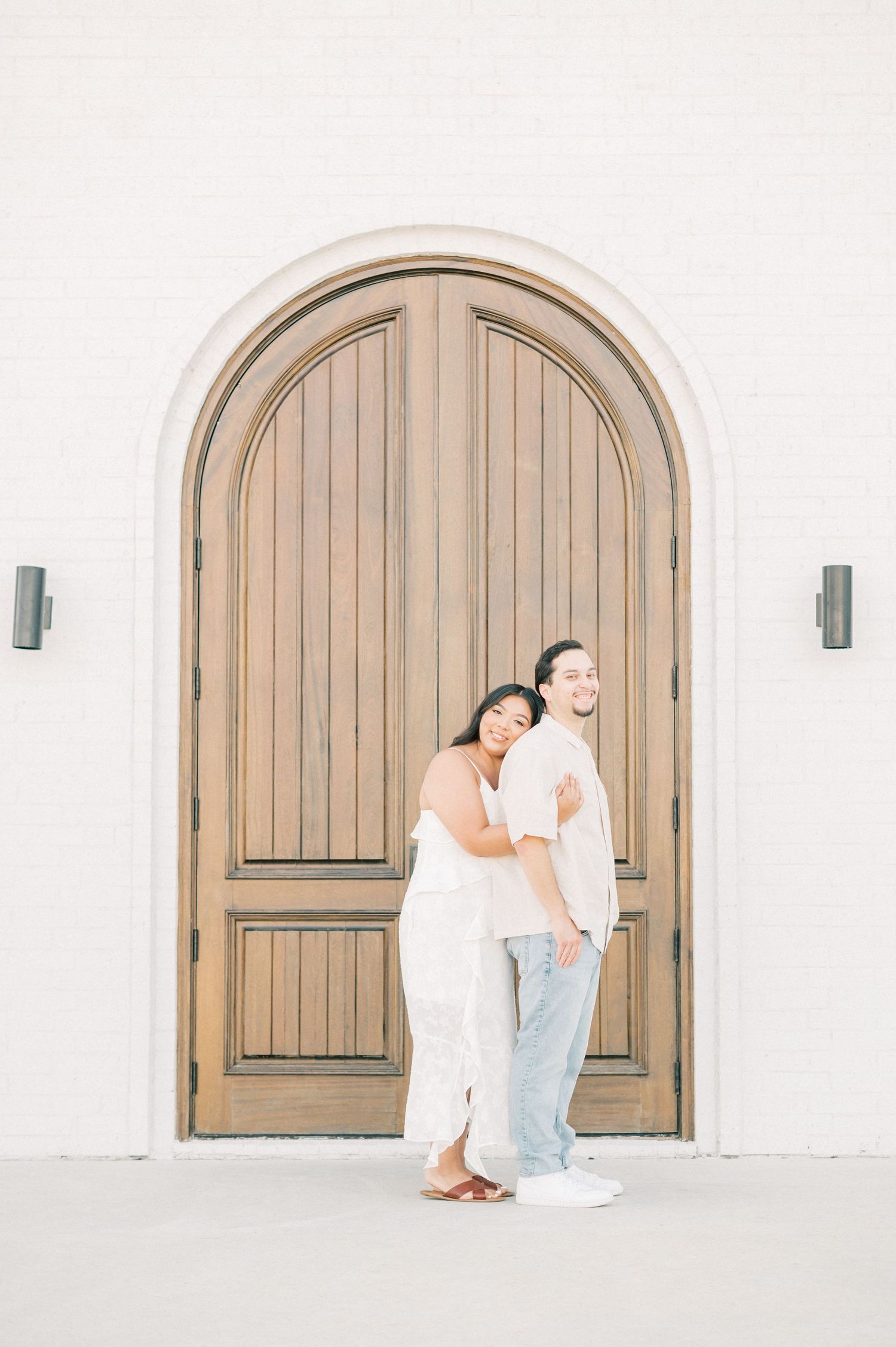 A couple embraces near a Mediterranean-style arched wooden doorway against a white building exterior.