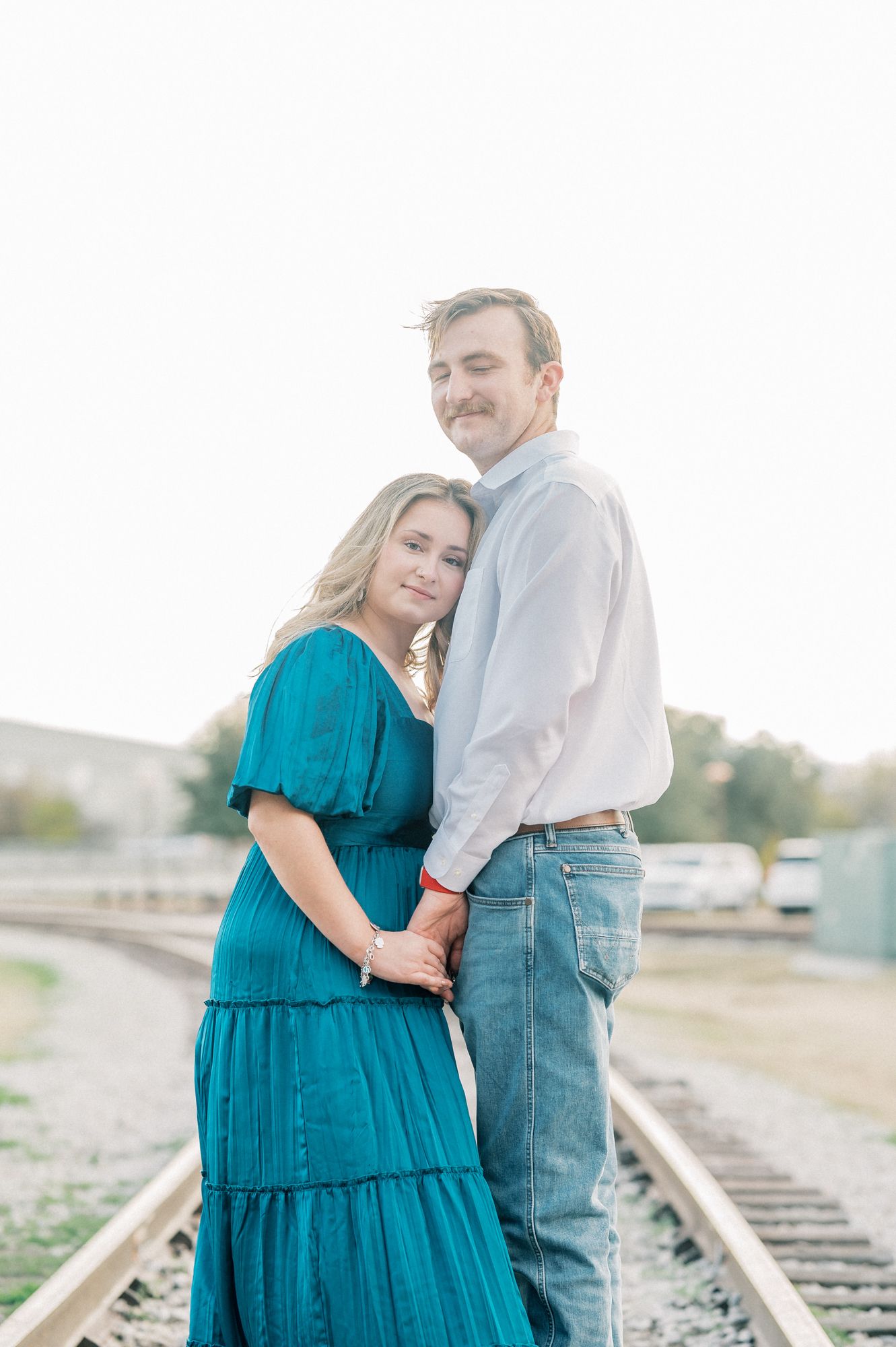 A romantic moment captured on train tracks as a couple in teal dress and white shirt embrace.