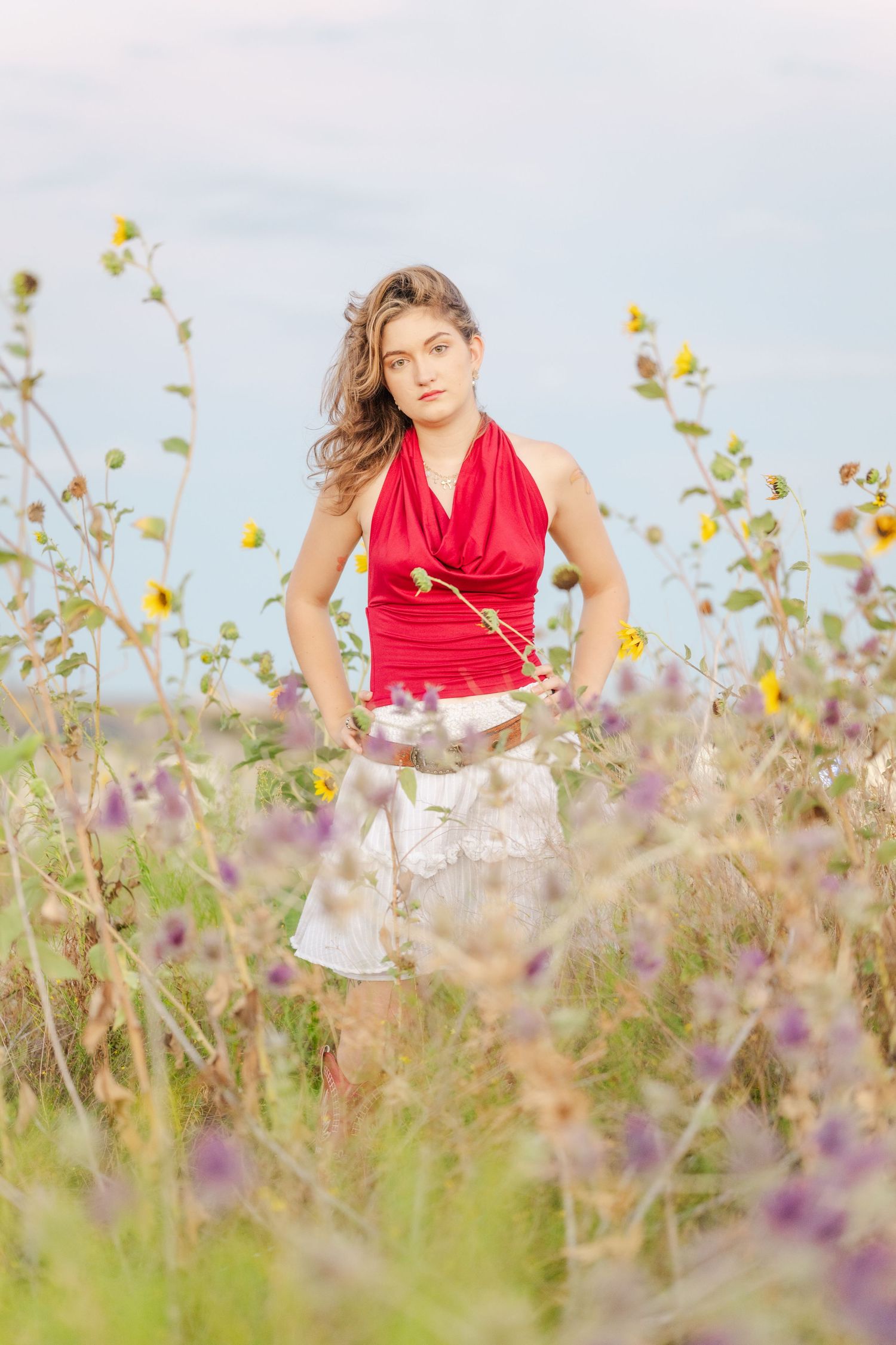 Woman in red halter top and white skirt standing in a wildflower field holding a sunflower on a cloudy day.