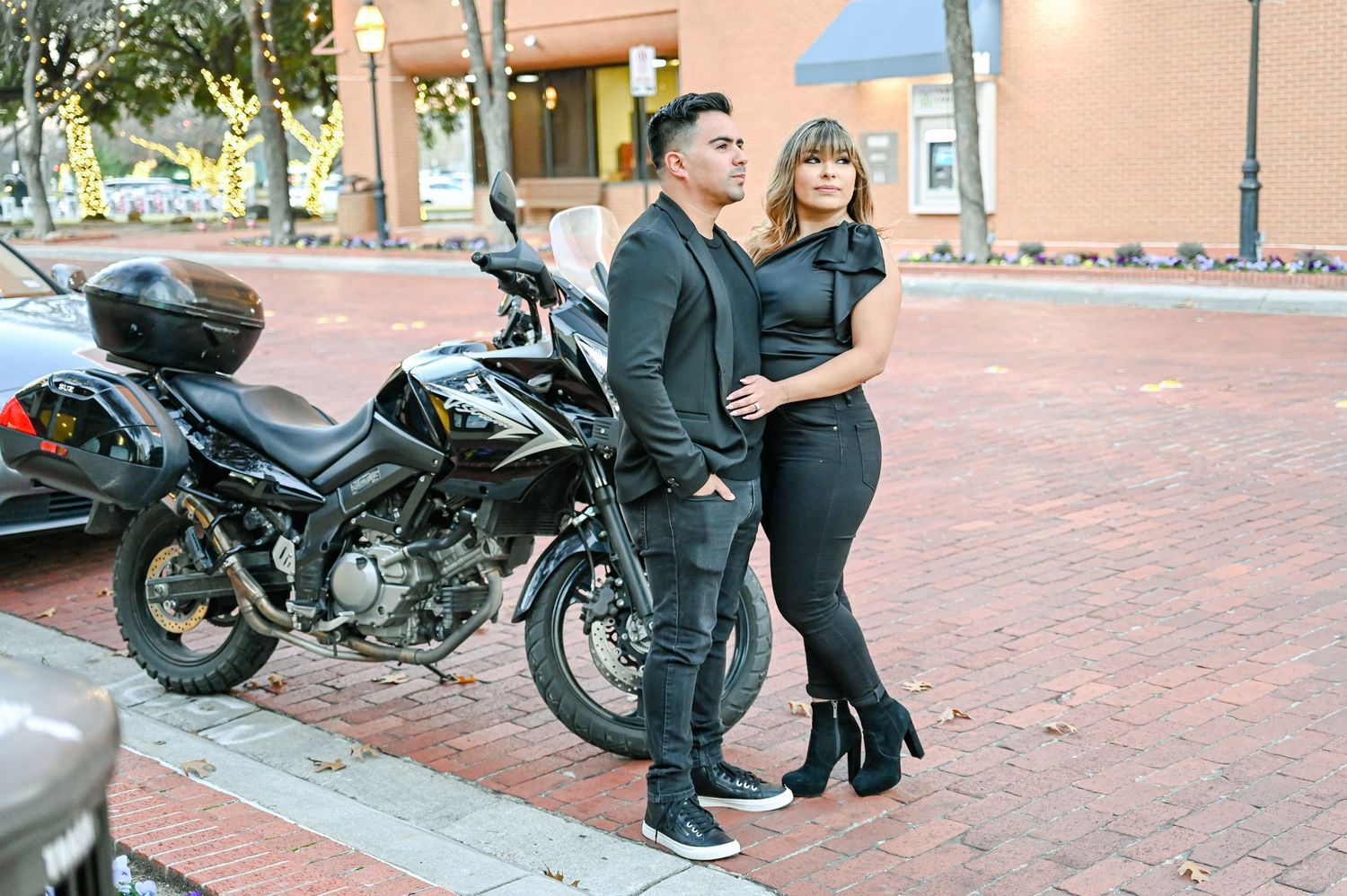 Couple in matching black outfits posing beside a black motorcycle on a brick sidewalk in an urban setting.
