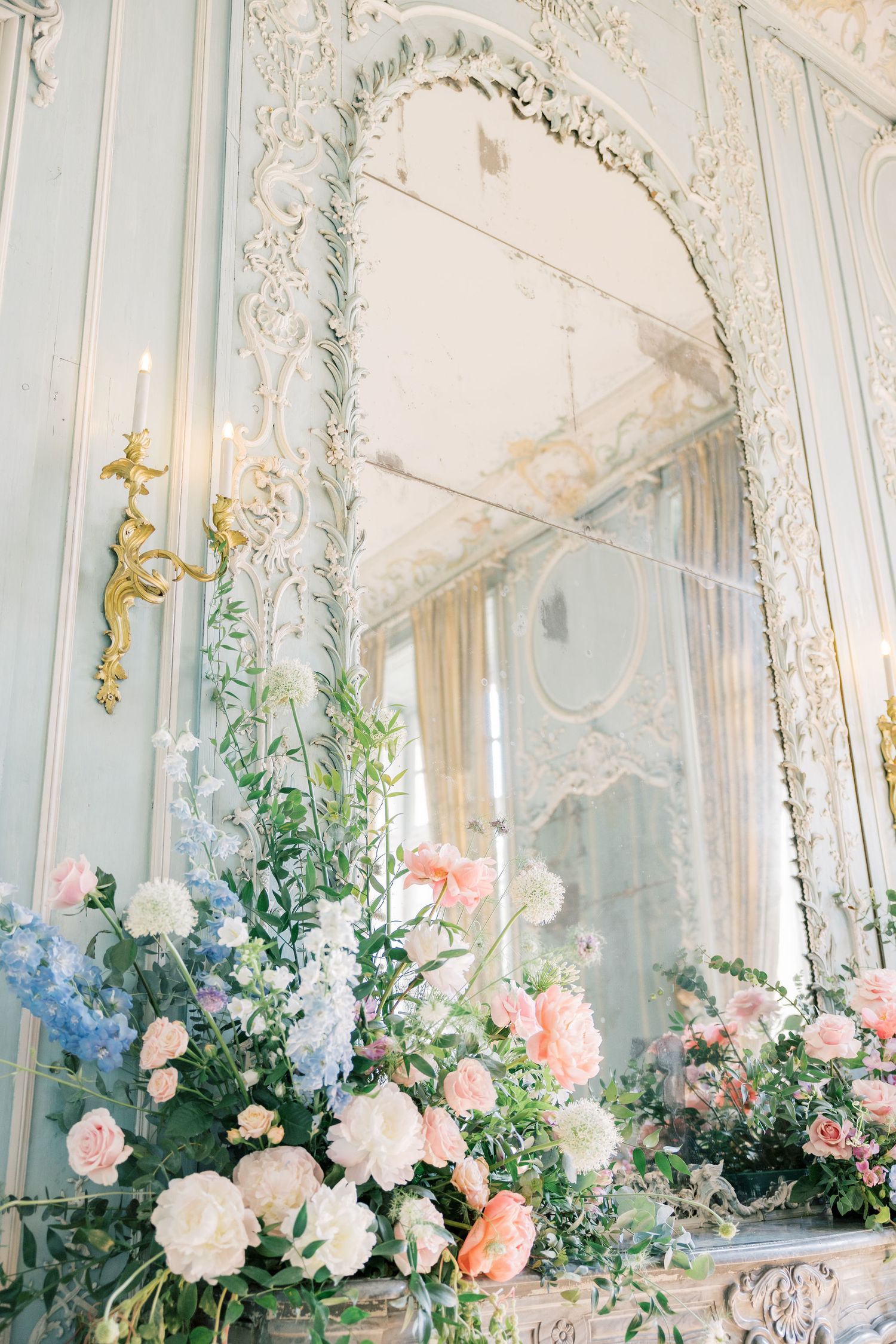 Ornate French baroque mirror above a fireplace mantel adorned with lush pink, white, and blue floral arrangements.