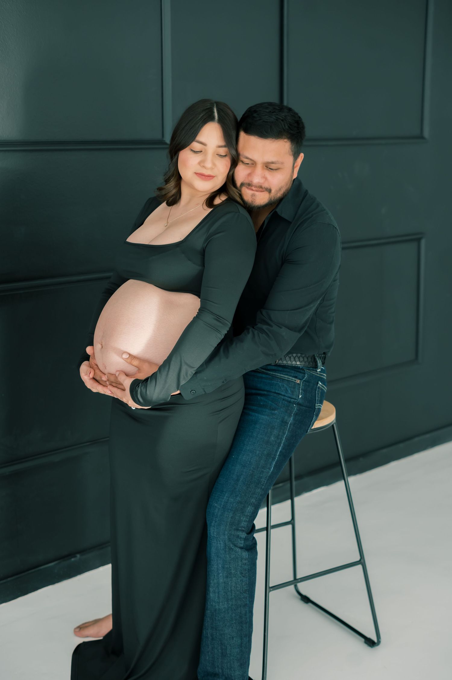 Couple shares tender moment during maternity session, man gently holding pregnant partners belly against dark green wall.