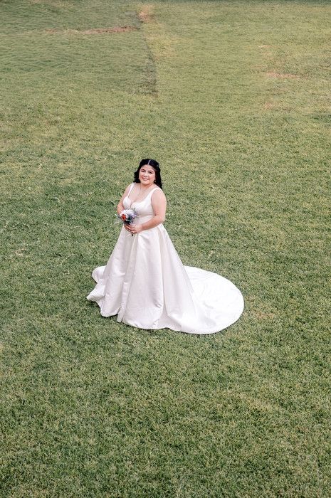 Bride in a white ball gown with train holds bouquet while standing on green grass lawn.