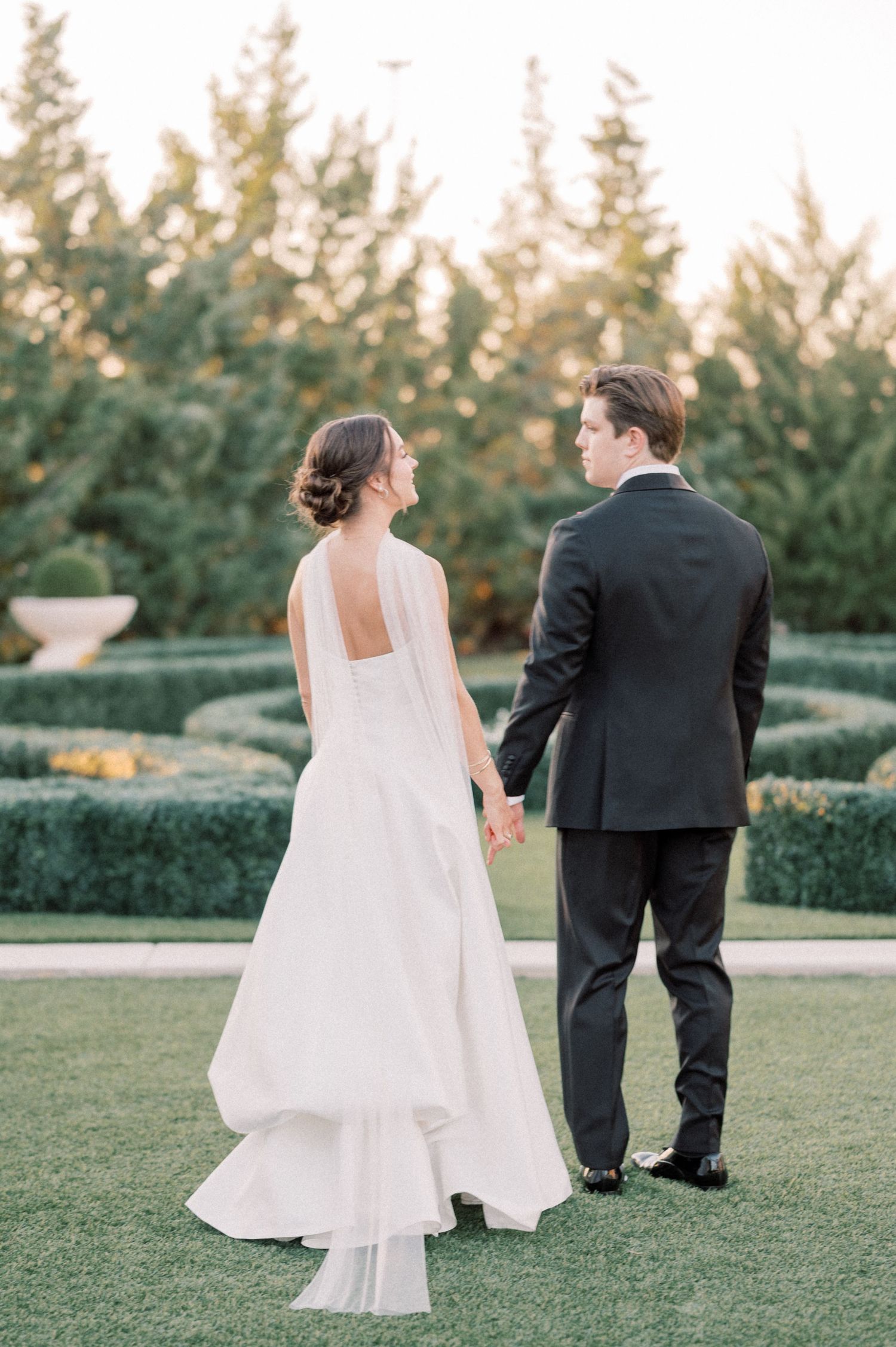 Bride and groom holding hands in a formal garden at golden hour, bride in elegant white gown with open back.