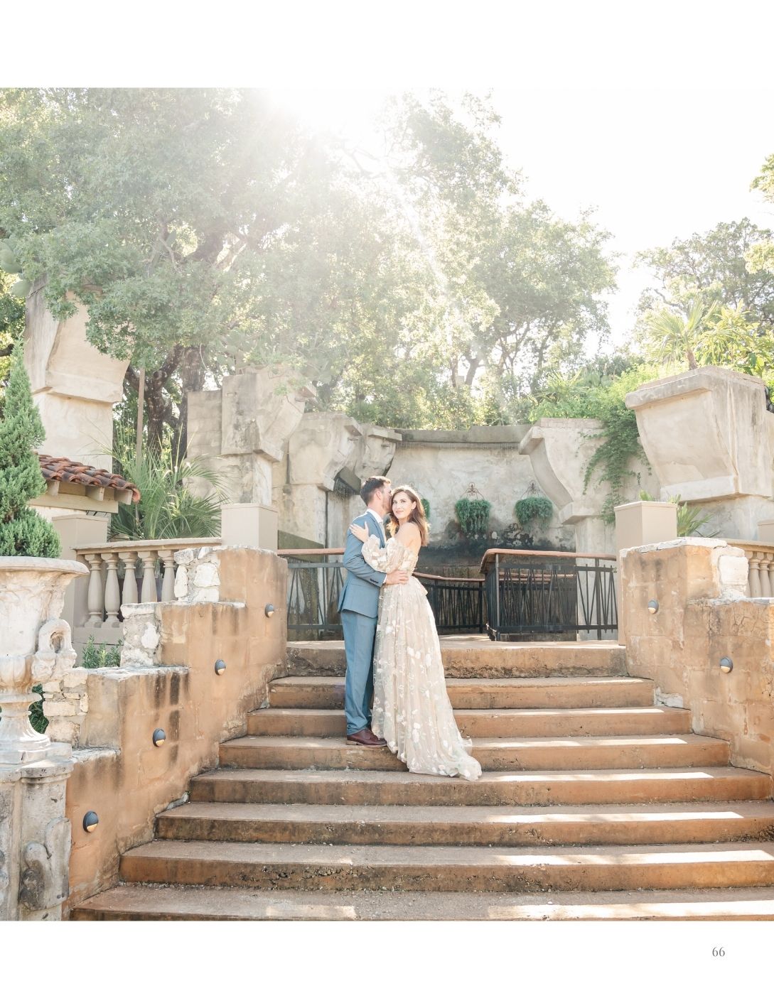 Couple embraces on stone steps at elegant outdoor wedding venue surrounded by lush trees and warm sunlight.