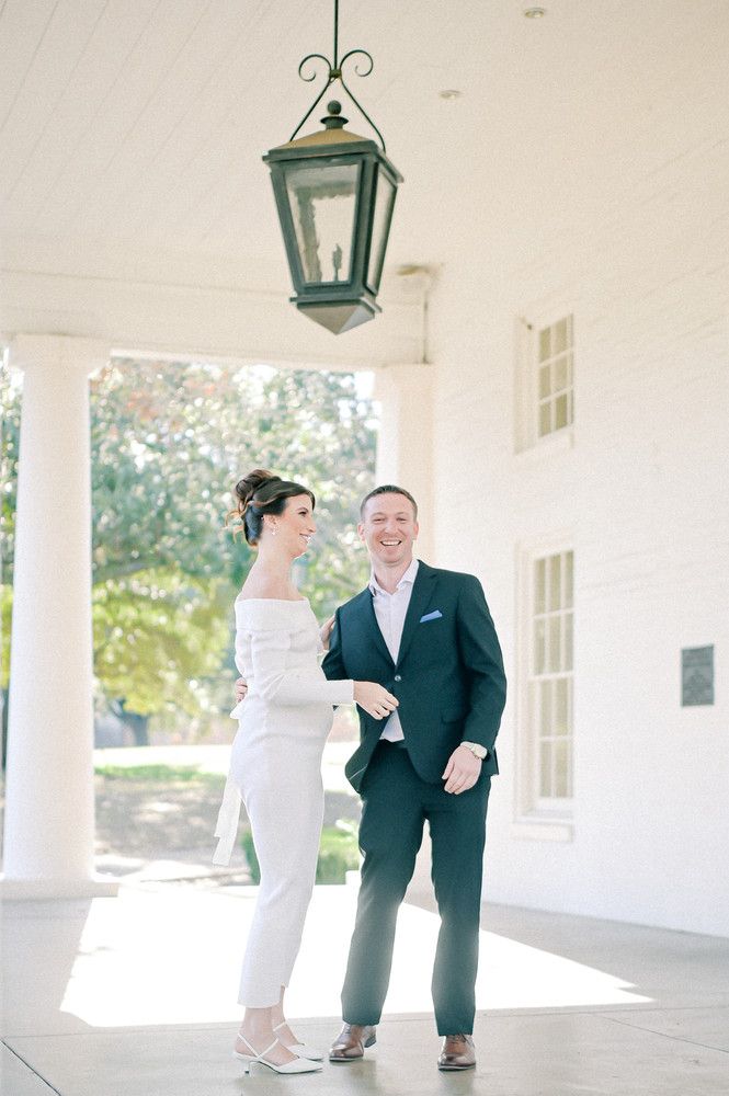 Elegant couple in white and navy attire laugh together on a sunlit colonial-style porch with lantern.