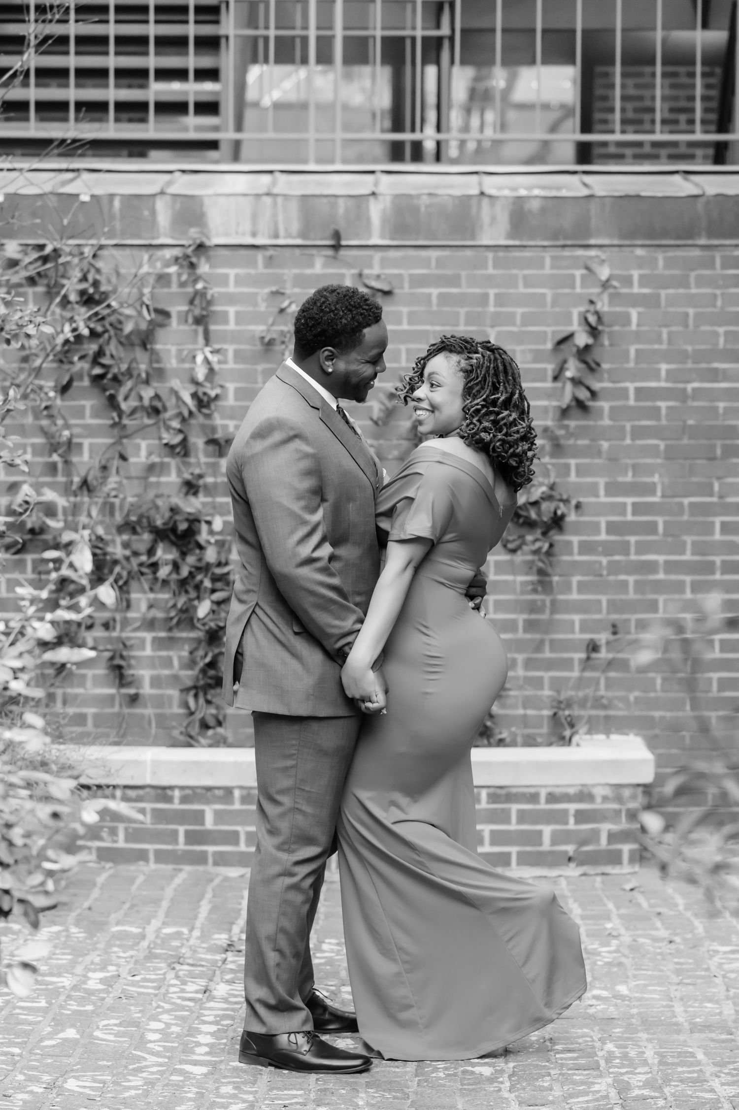 Black and white photo of a couple in formal wear embracing tenderly against a brick wall with ivy.
