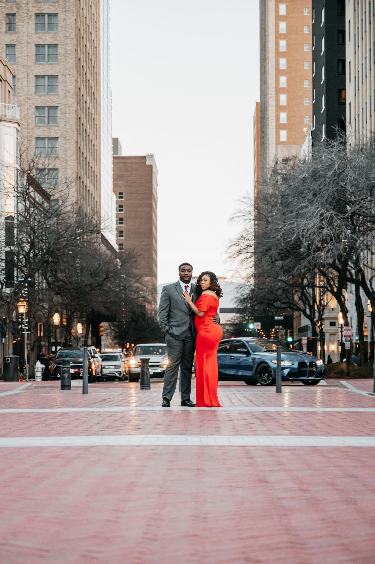 Engaged couple in red dress and gray suit posing on urban city street at dusk.