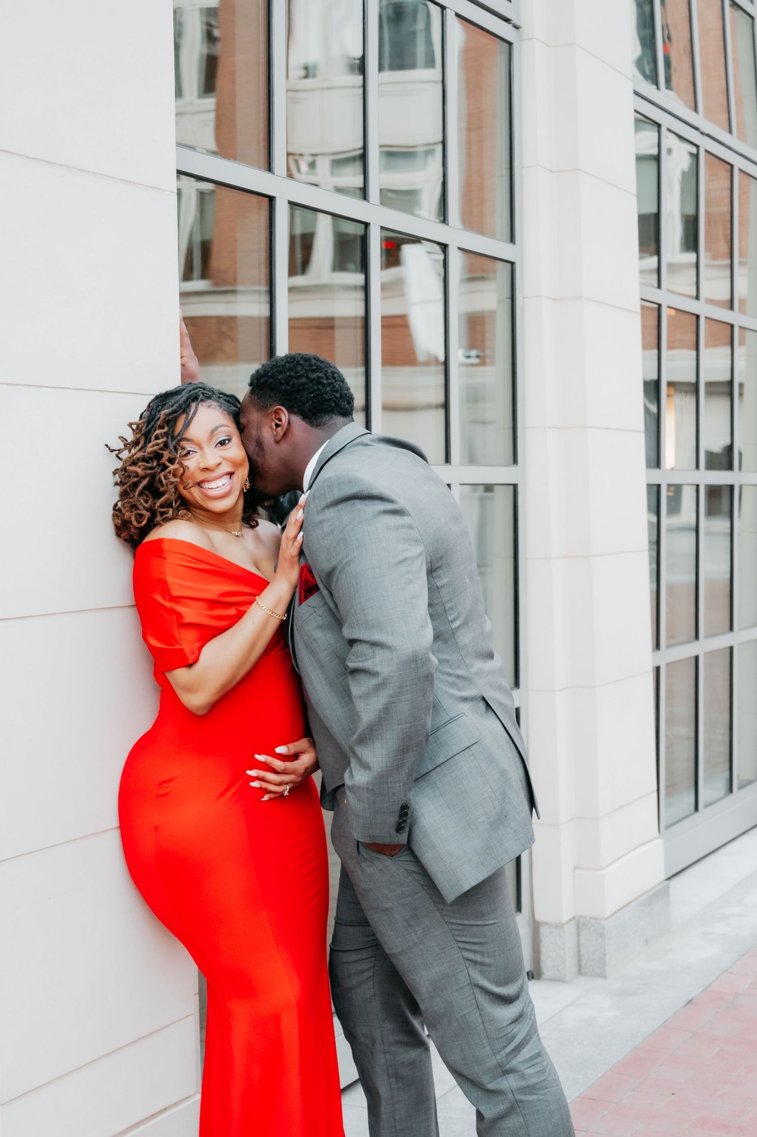 A couple poses romantically outside, woman in red gown, man in gray suit kissing her cheek.