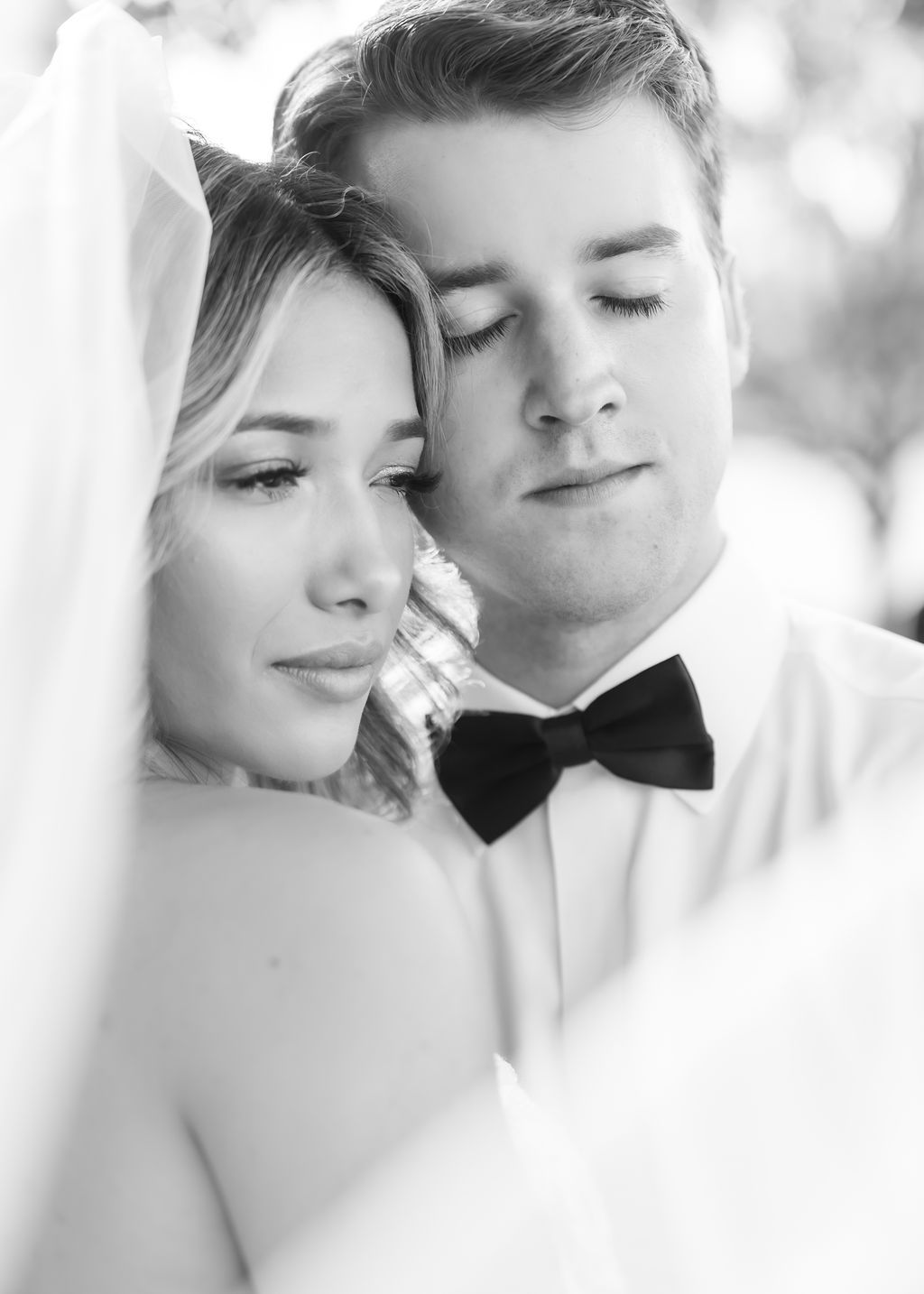Romantic black and white close-up portrait of bride and groom embracing tenderly under flowing wedding veil.