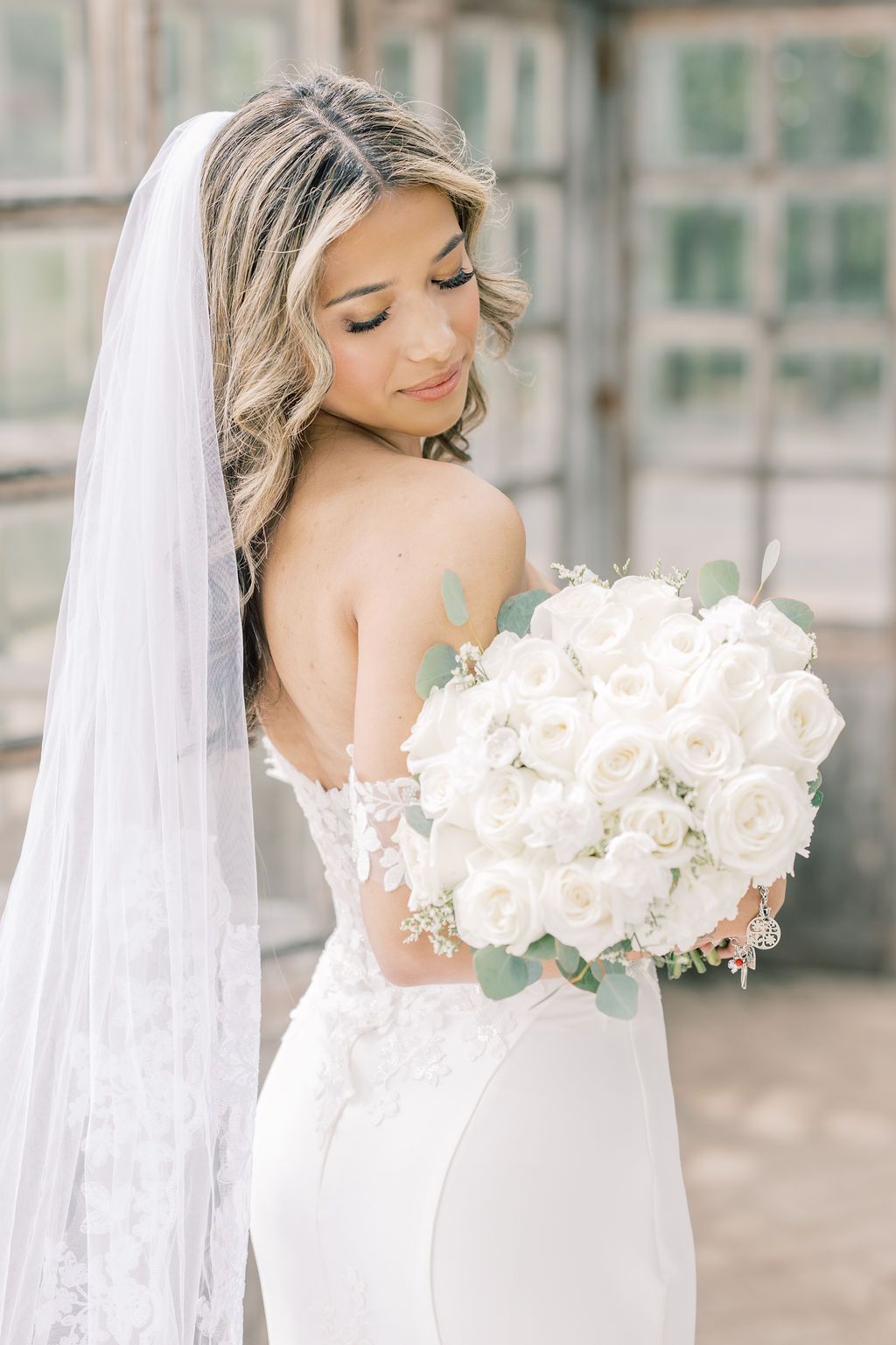 Bride in lace backless gown holds white rose bouquet with veil cascading down her back.