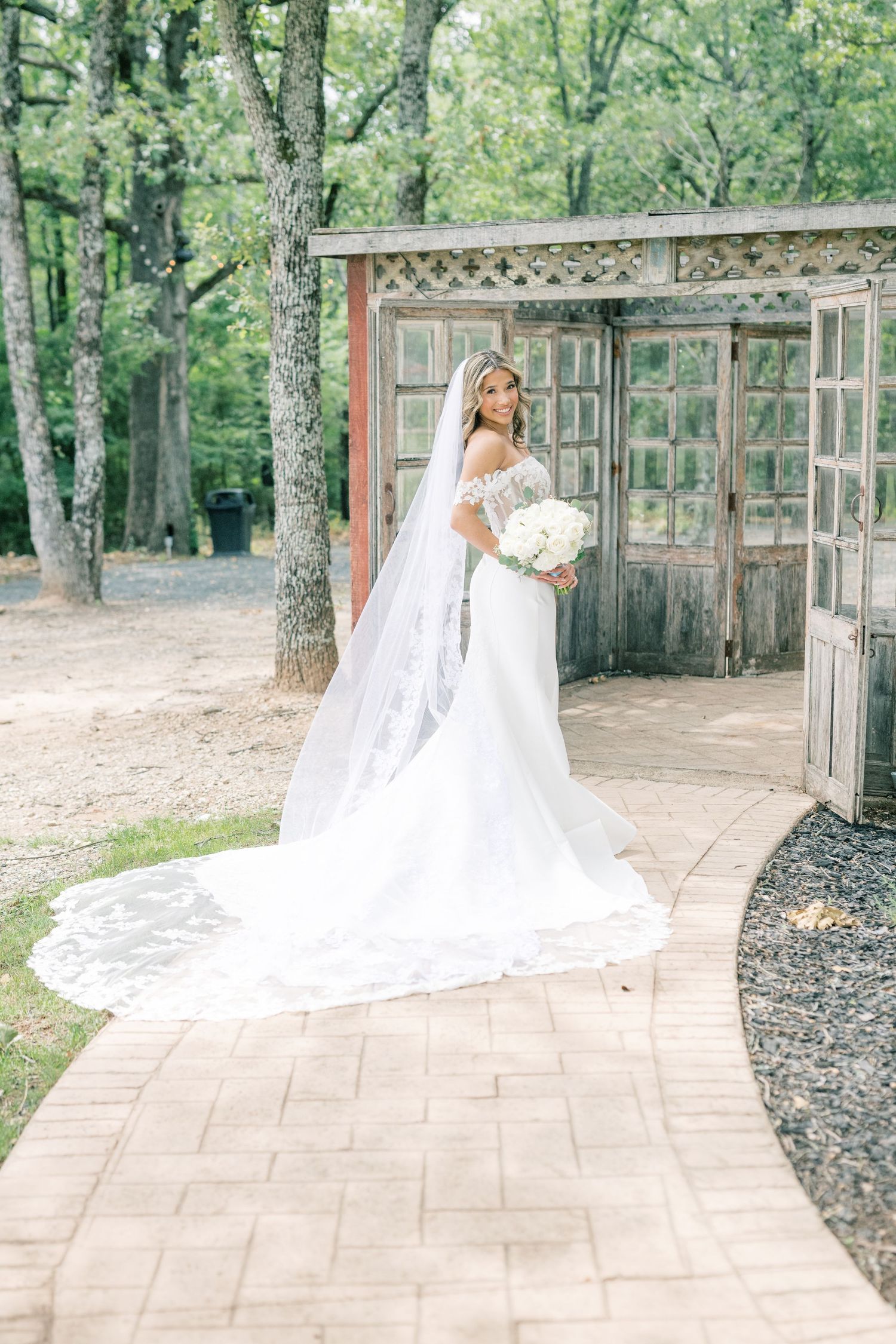 Bride in off-shoulder lace gown with cathedral veil poses on brick path near rustic wooden doors.
