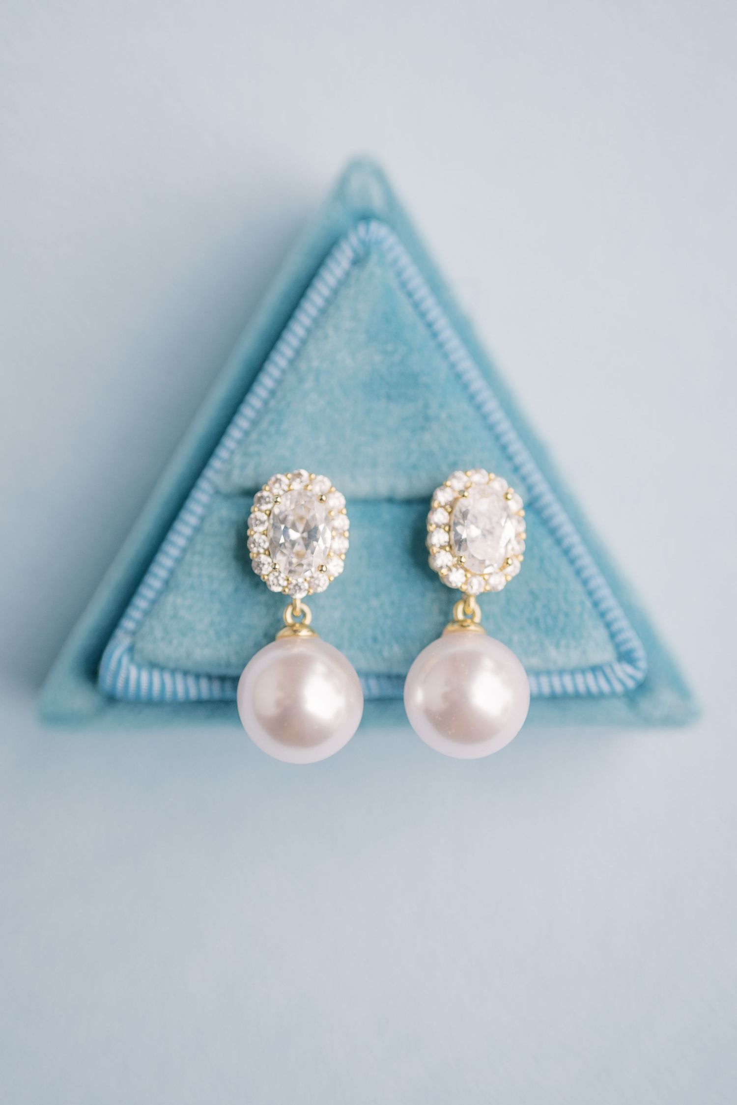 Elegant pearl drop earrings with crystal halo studs in gold setting displayed in a blue velvet triangular jewelry box.
