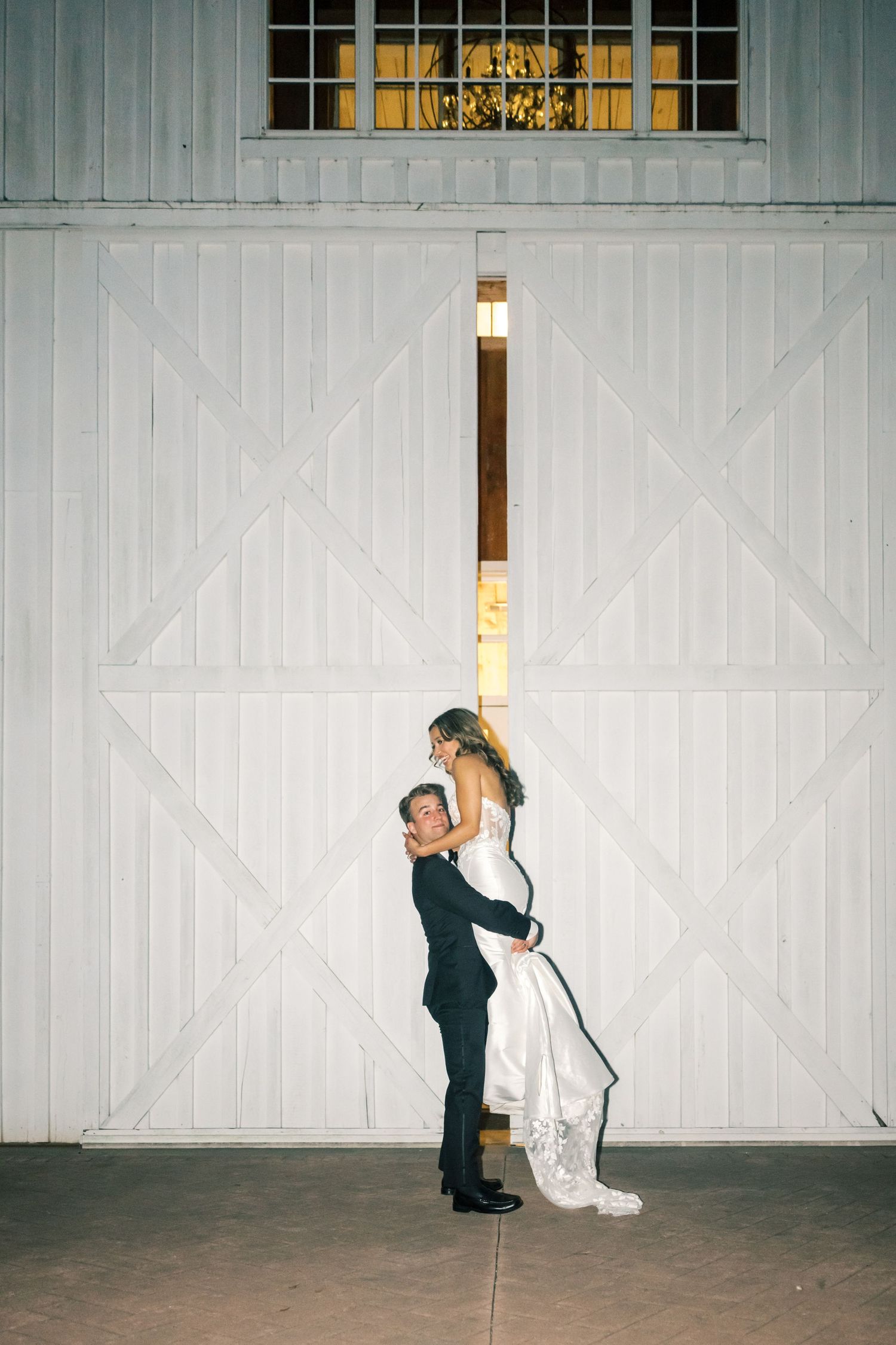 Groom lifting bride in front of white barn doors during romantic nighttime wedding portrait.