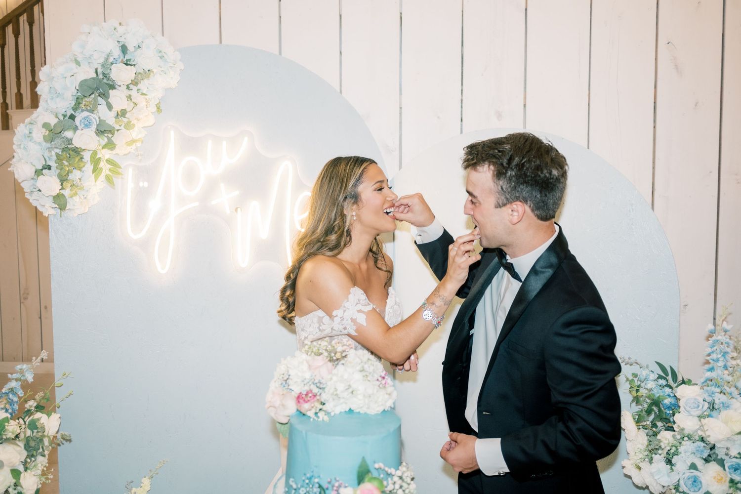 Bride and groom sharing cake at reception in front of a blue floral backdrop with neon sign.