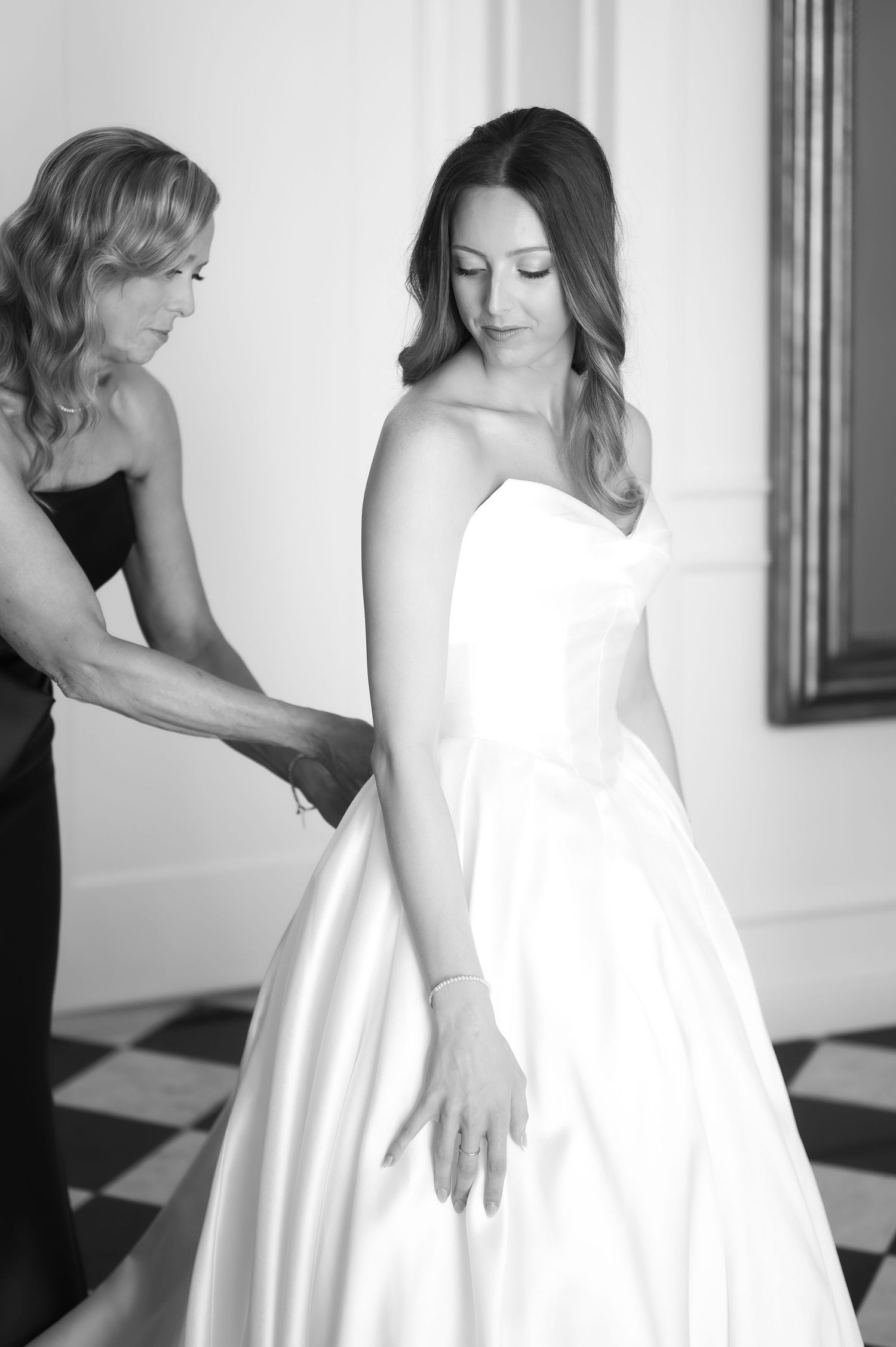 Black and white photo of a bride in a strapless gown being helped by a woman in a dark dress.