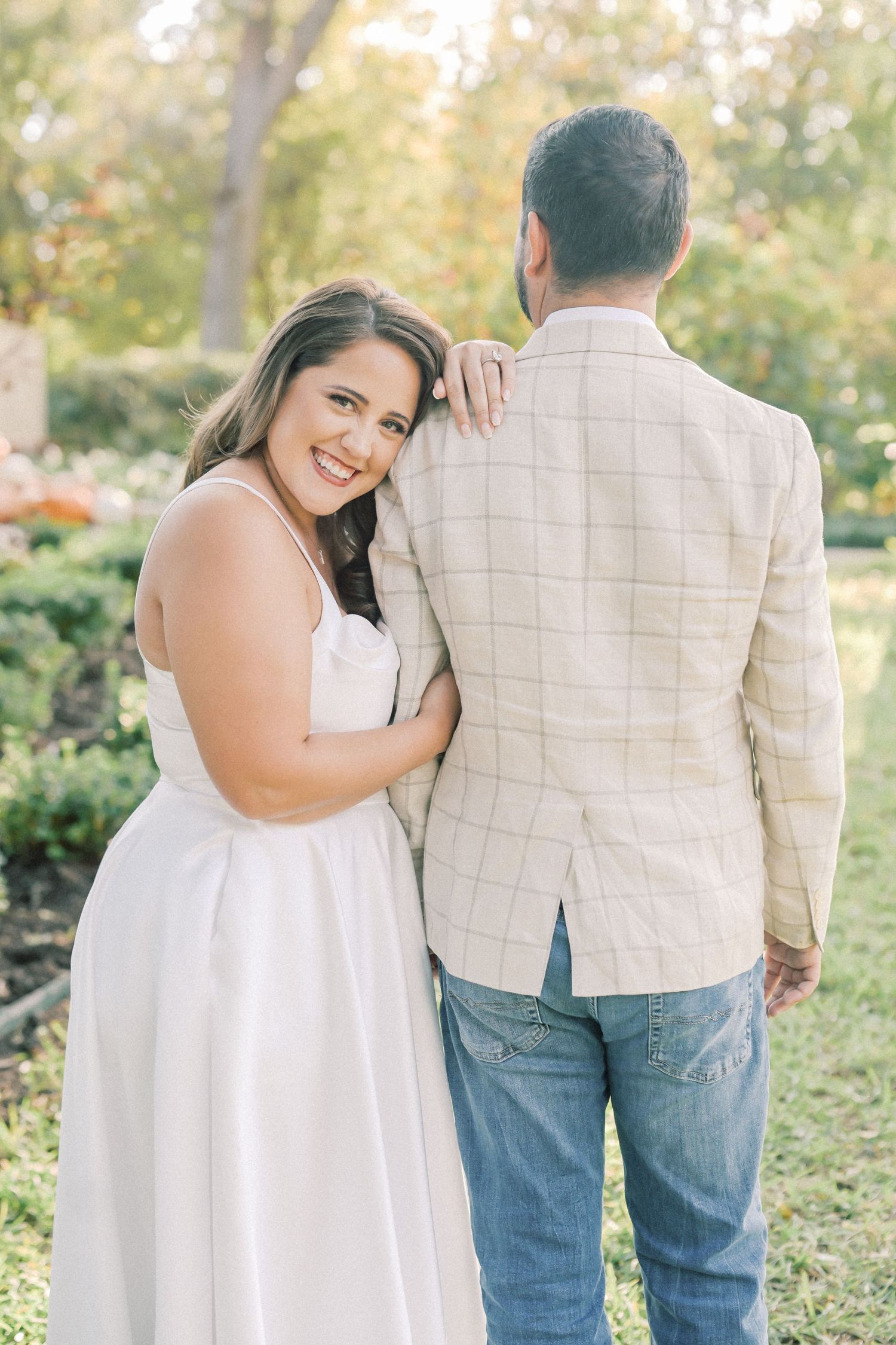 Smiling woman in a white dress looks over her shoulder while hugging her fiancé from the side in a lush garden engagement session.