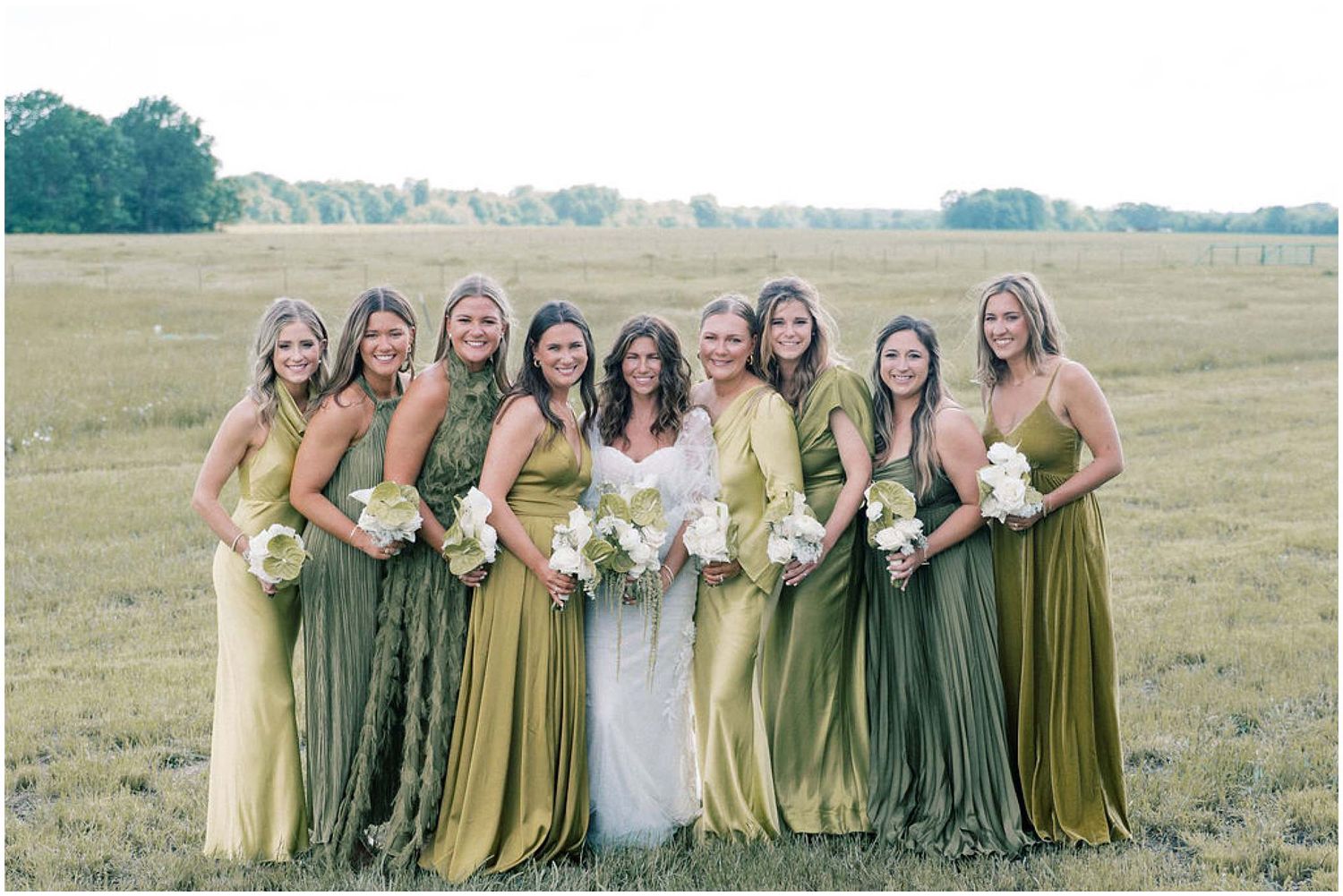 Bridesmaids in varying shades of green and yellow dresses pose together in an open field.