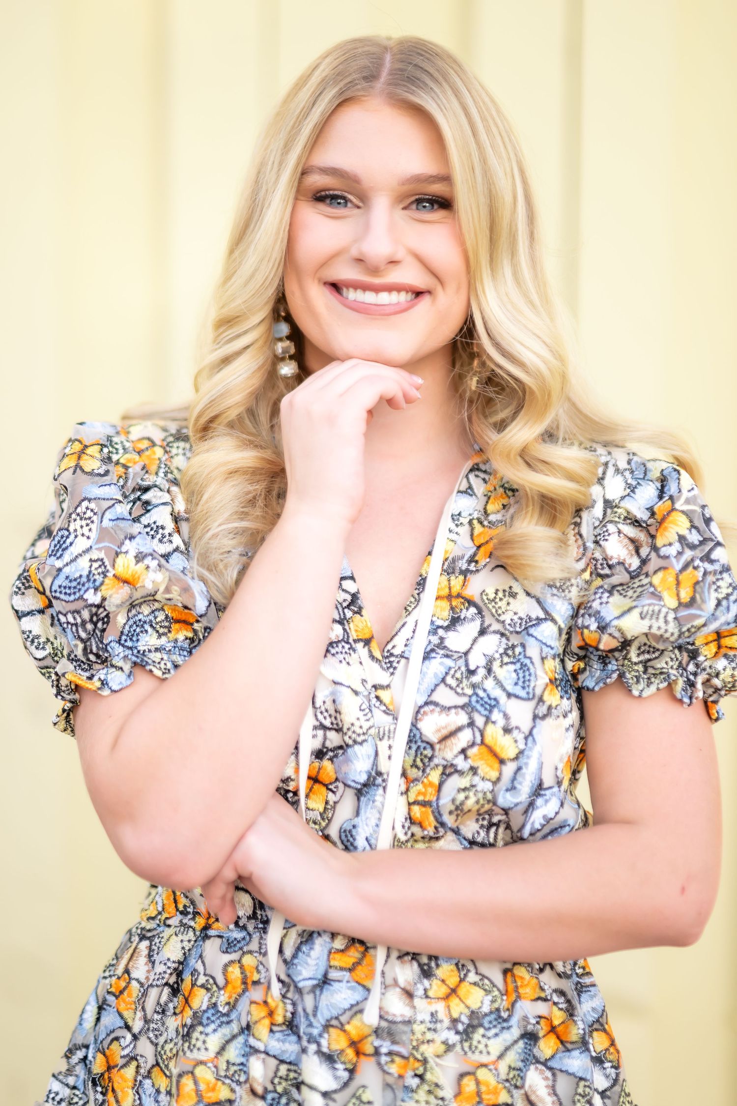 Smiling blonde woman in floral dress with butterfly print posing confidently against a soft yellow background.