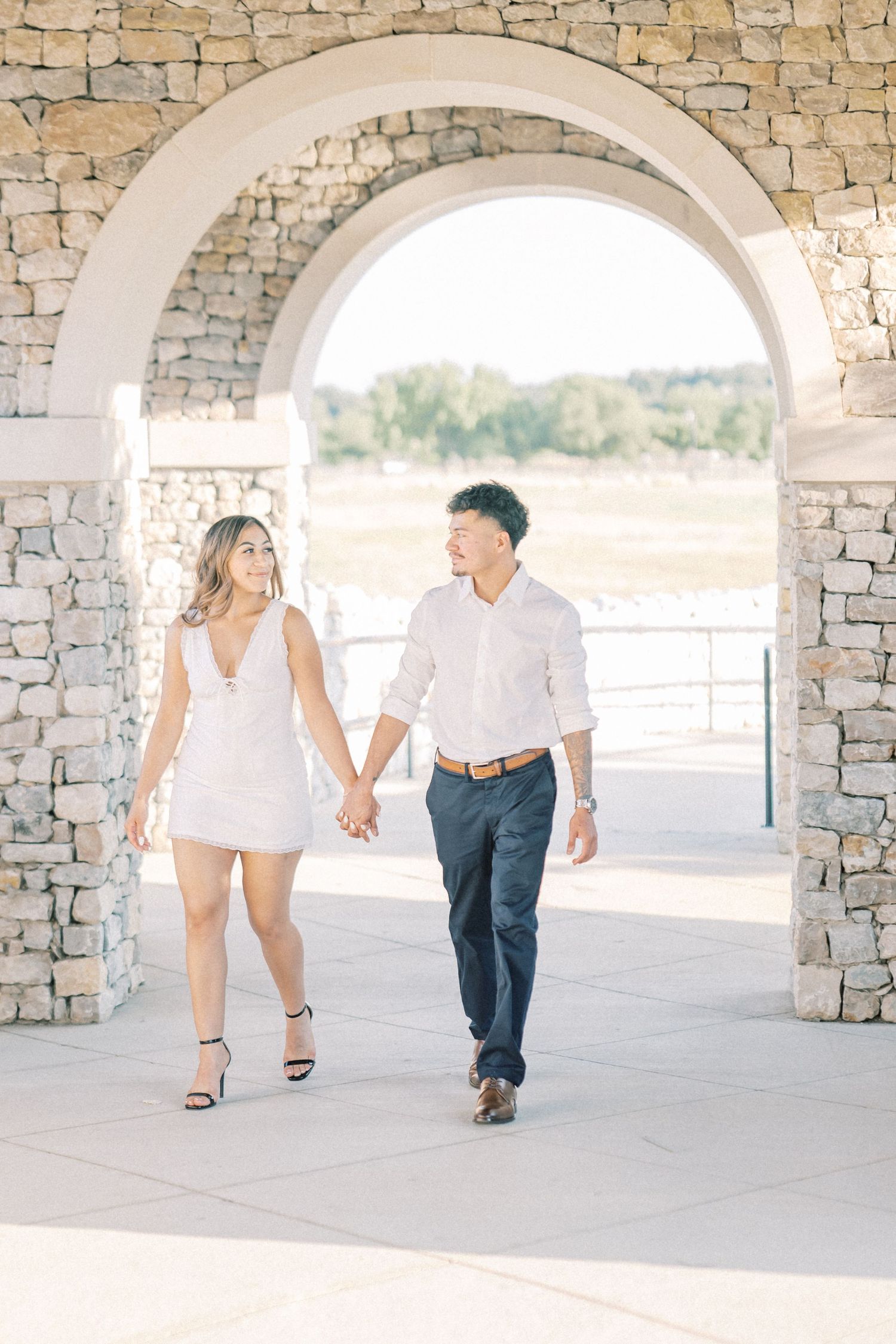 Couple holding hands walking through stone arch during engagement photo session.