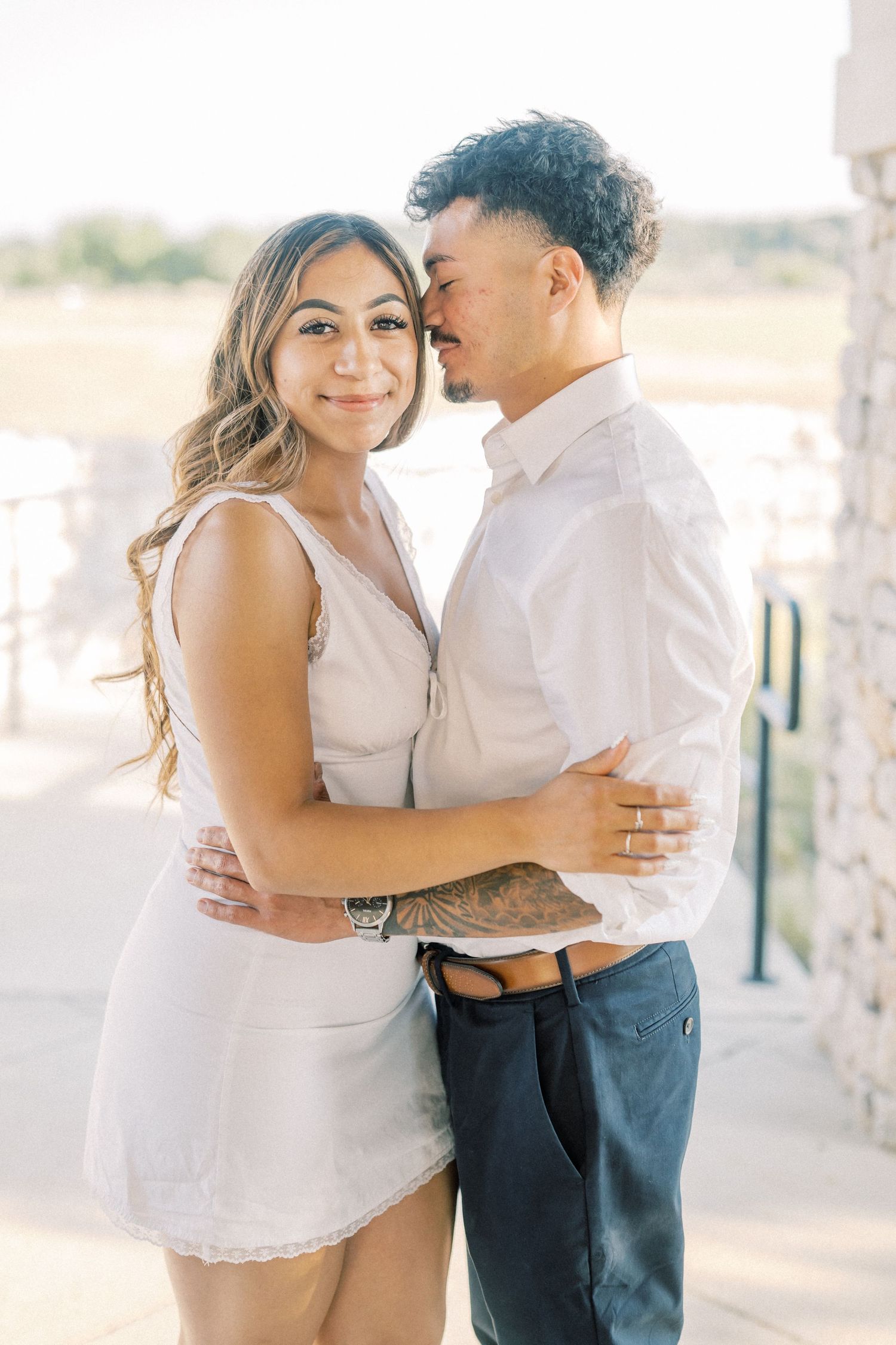 Couple embracing outdoors, woman in white dress smiling at camera while man kisses her cheek, soft natural lighting.