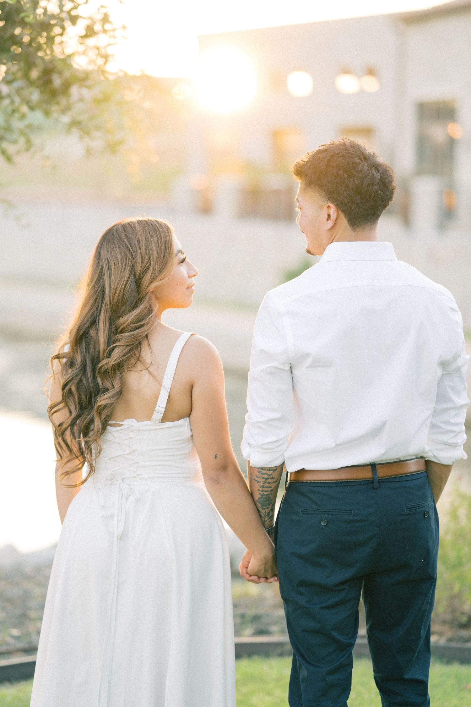 An engaged couple holds hands while walking away from the camera, bathed in warm sunset light.