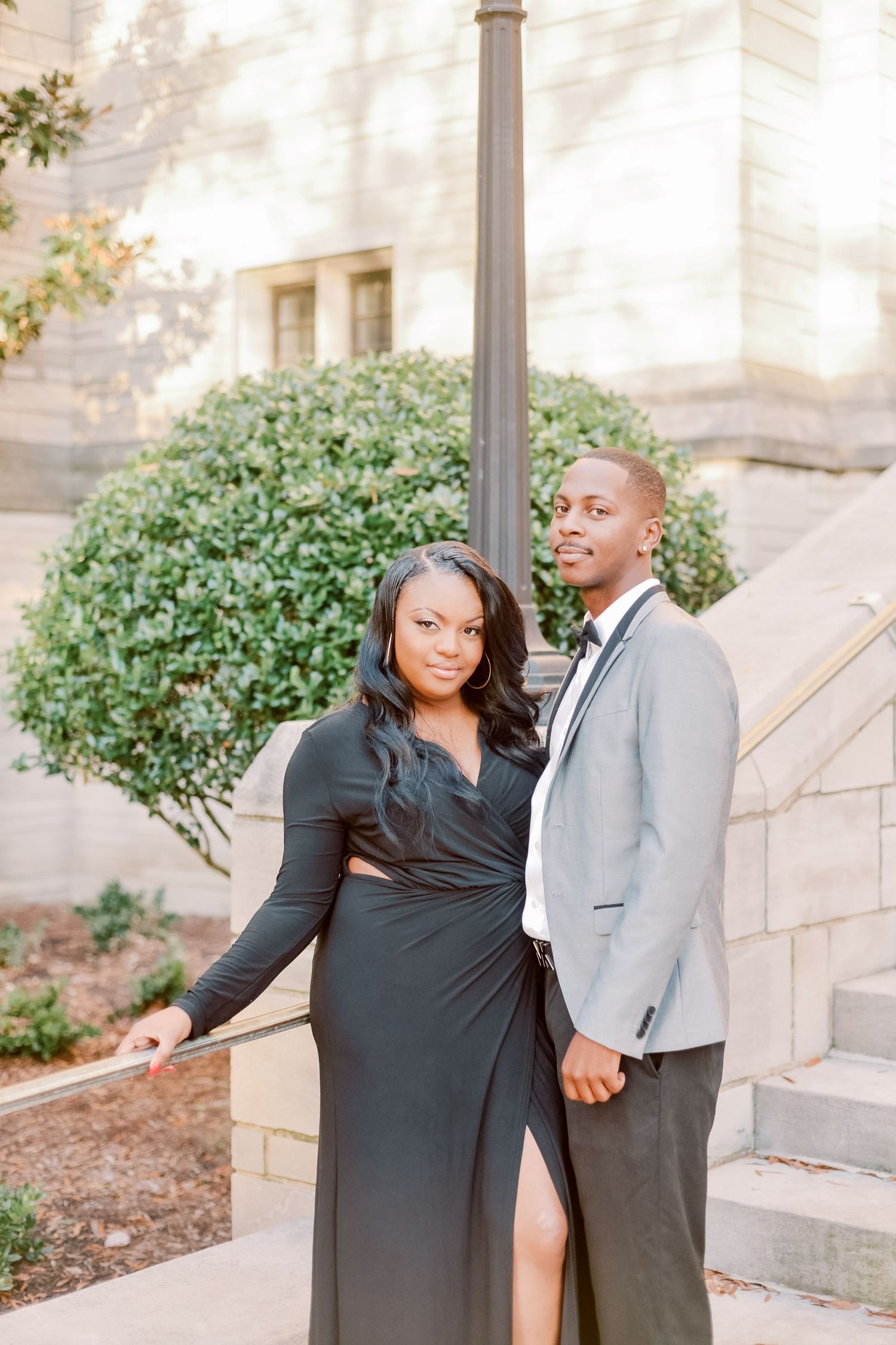 Elegant couple posing outdoors, woman in black wrap dress and man in gray suit, standing near a lamppost.