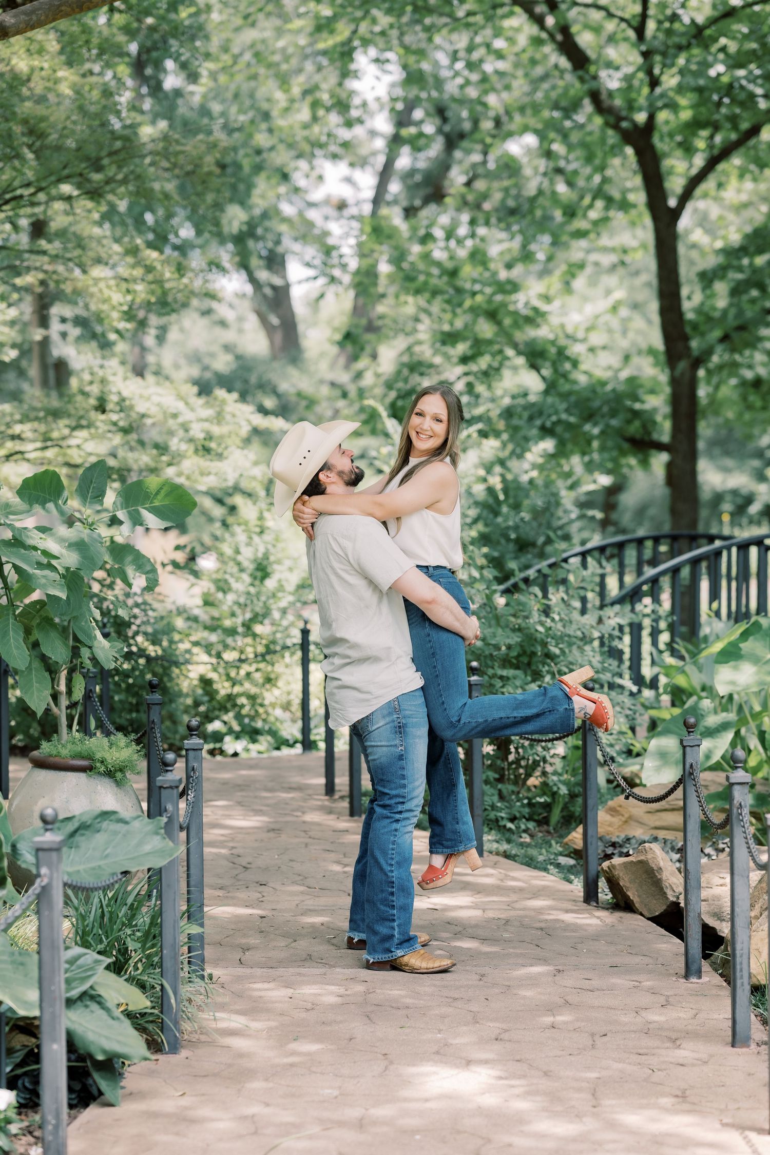 Man lifting woman on a garden bridge path surrounded by greenery.