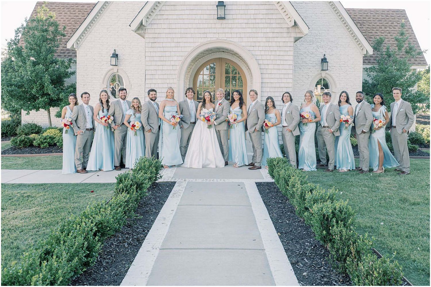 Large wedding party poses outside a stone chapel, bridesmaids in sage green gowns and groomsmen in gray suits.