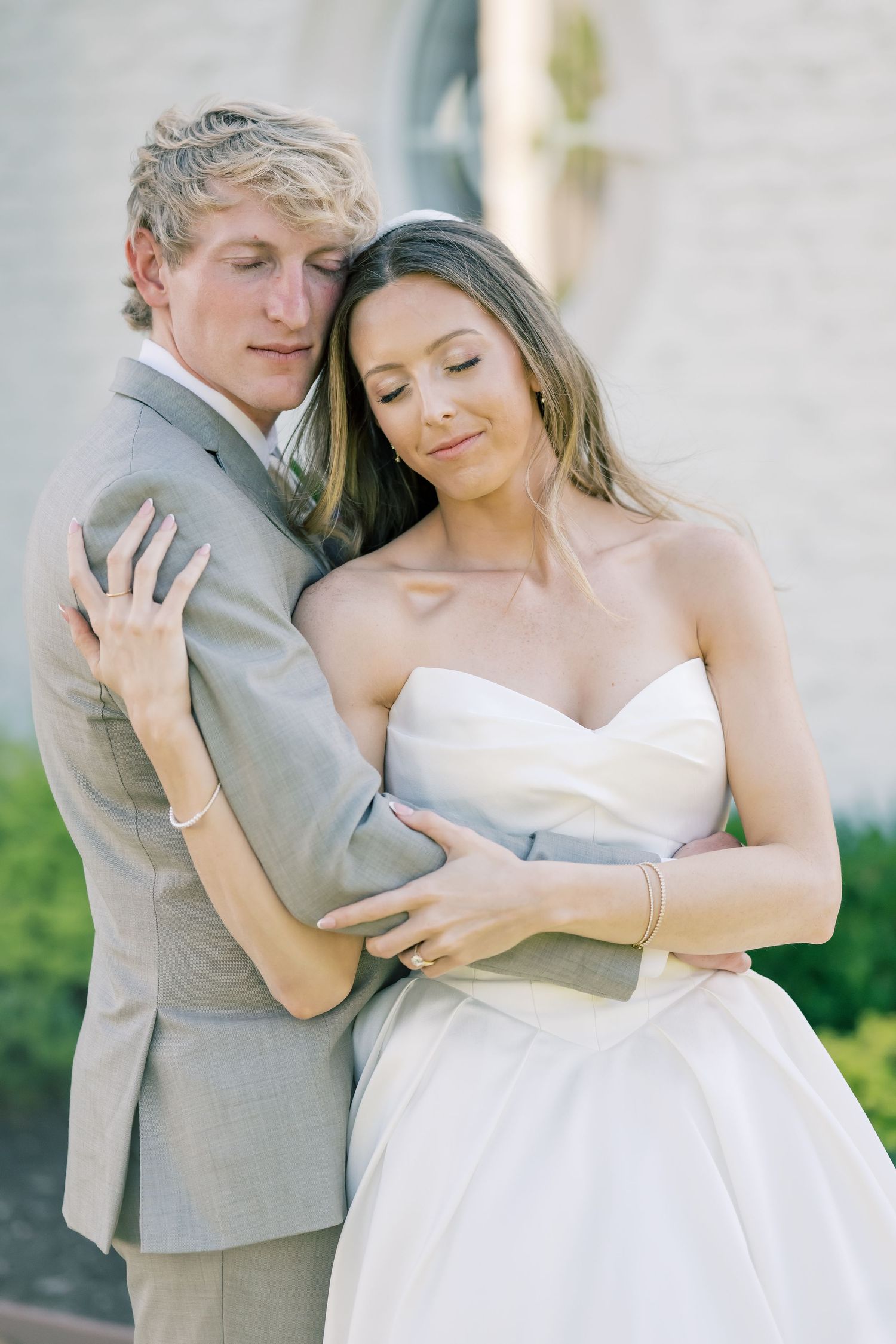 Bride and groom in tender embrace outdoors, groom in gray suit, bride in strapless gown.