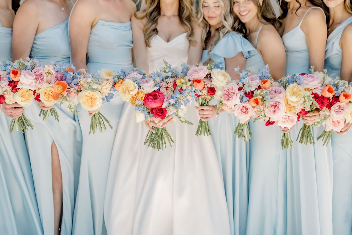 Bride and bridesmaids in light blue dresses holding colorful floral bouquets with pink, red, and blue flowers.