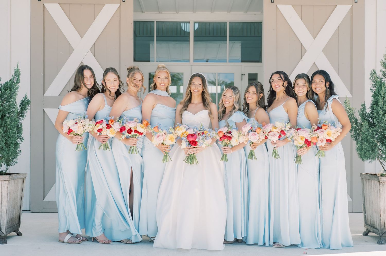 Bride in white gown with bridesmaids in light blue dresses holding colorful bouquets outside a white barn wedding venue.