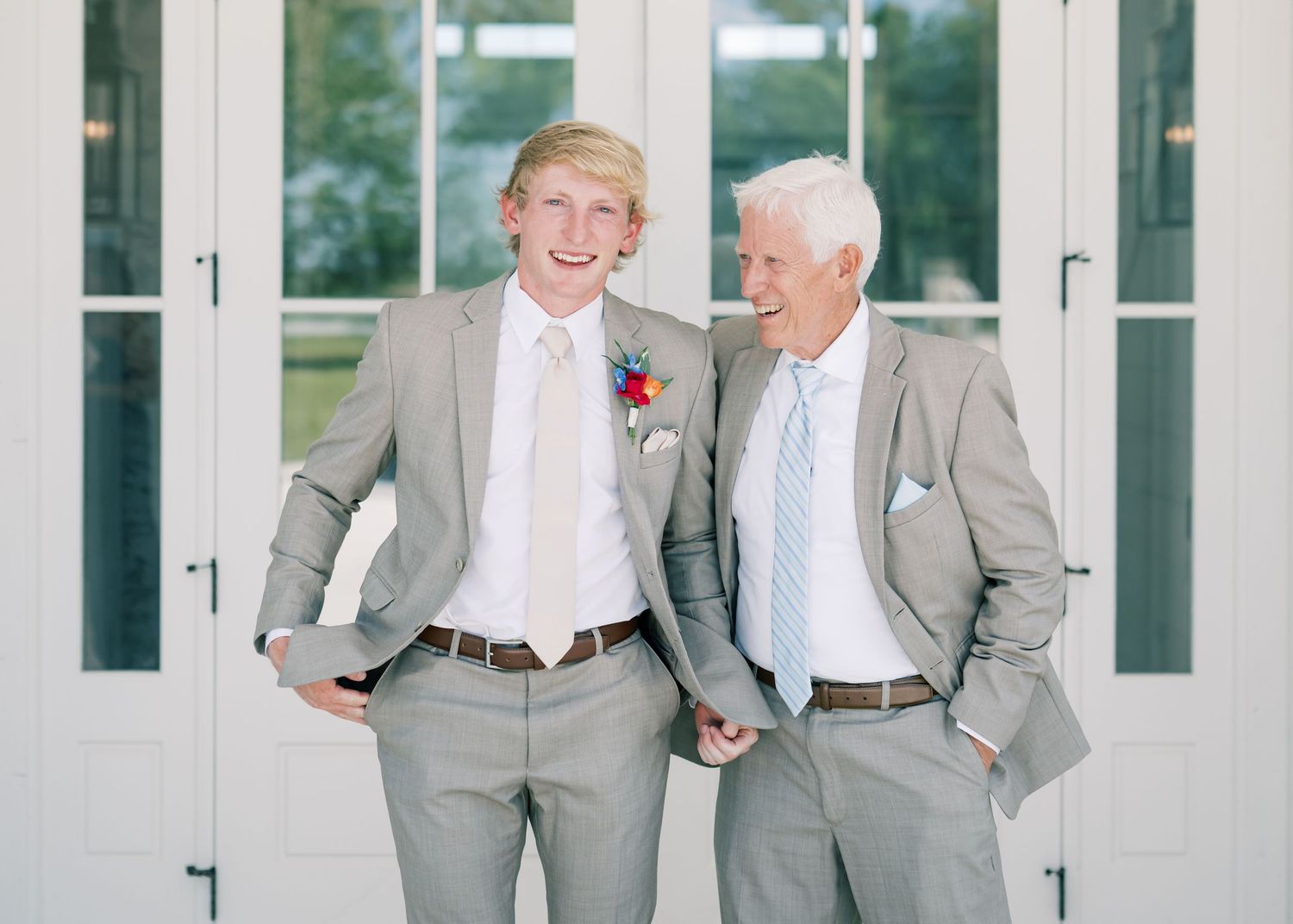 Two men in matching gray suits smile together outside white French doors at a wedding celebration.