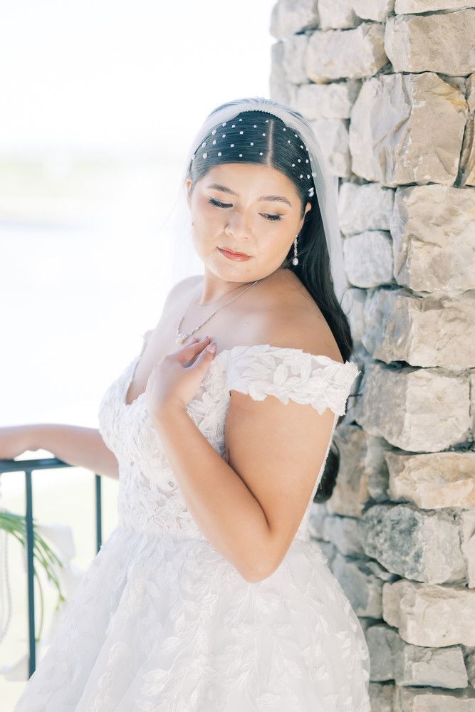 A bride in an off-shoulder gown and pearl headpiece poses beside a stone column.