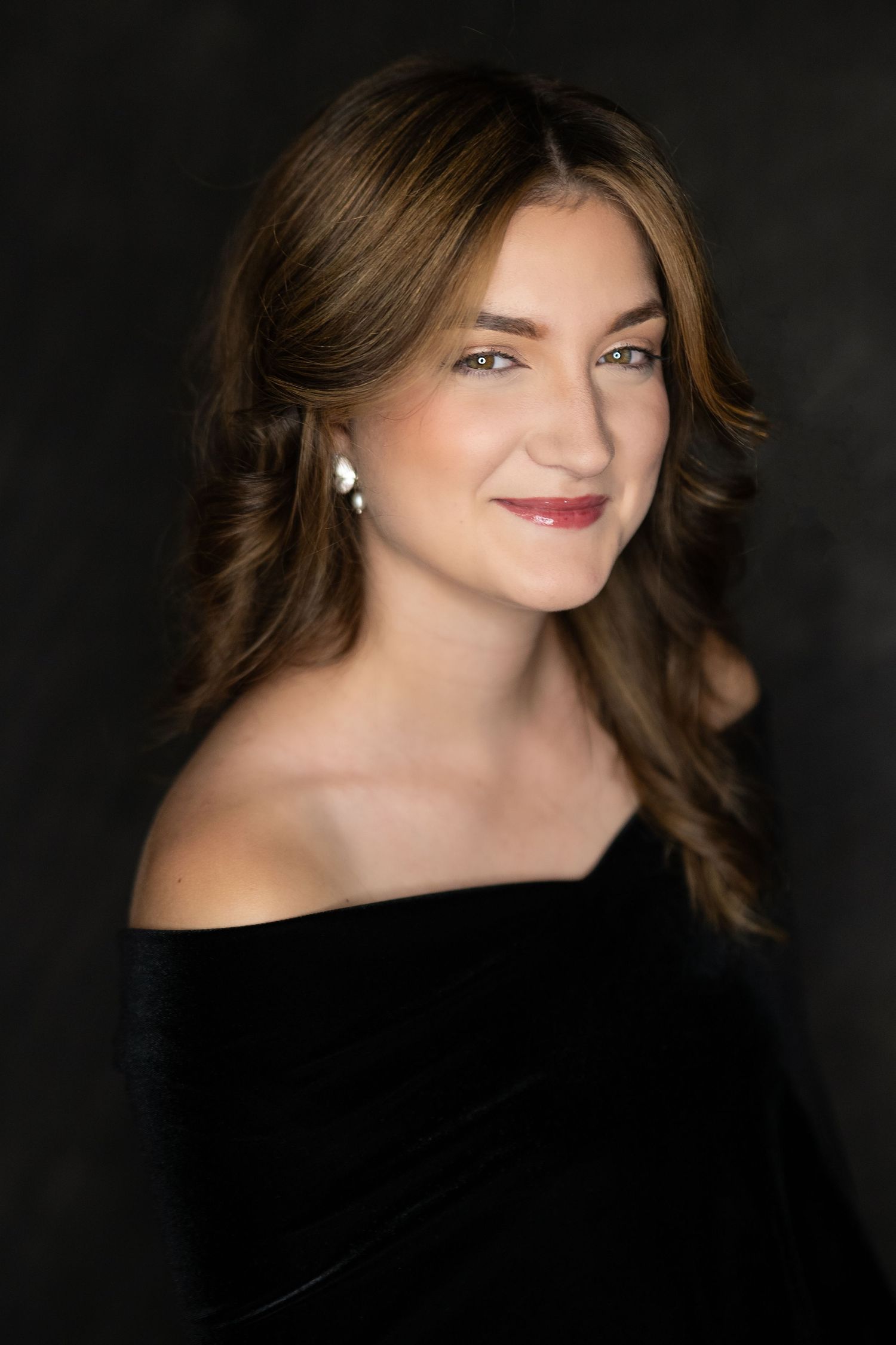 Woman with brown hair smiling in black off-shoulder top, wearing pearl earrings against dark background.