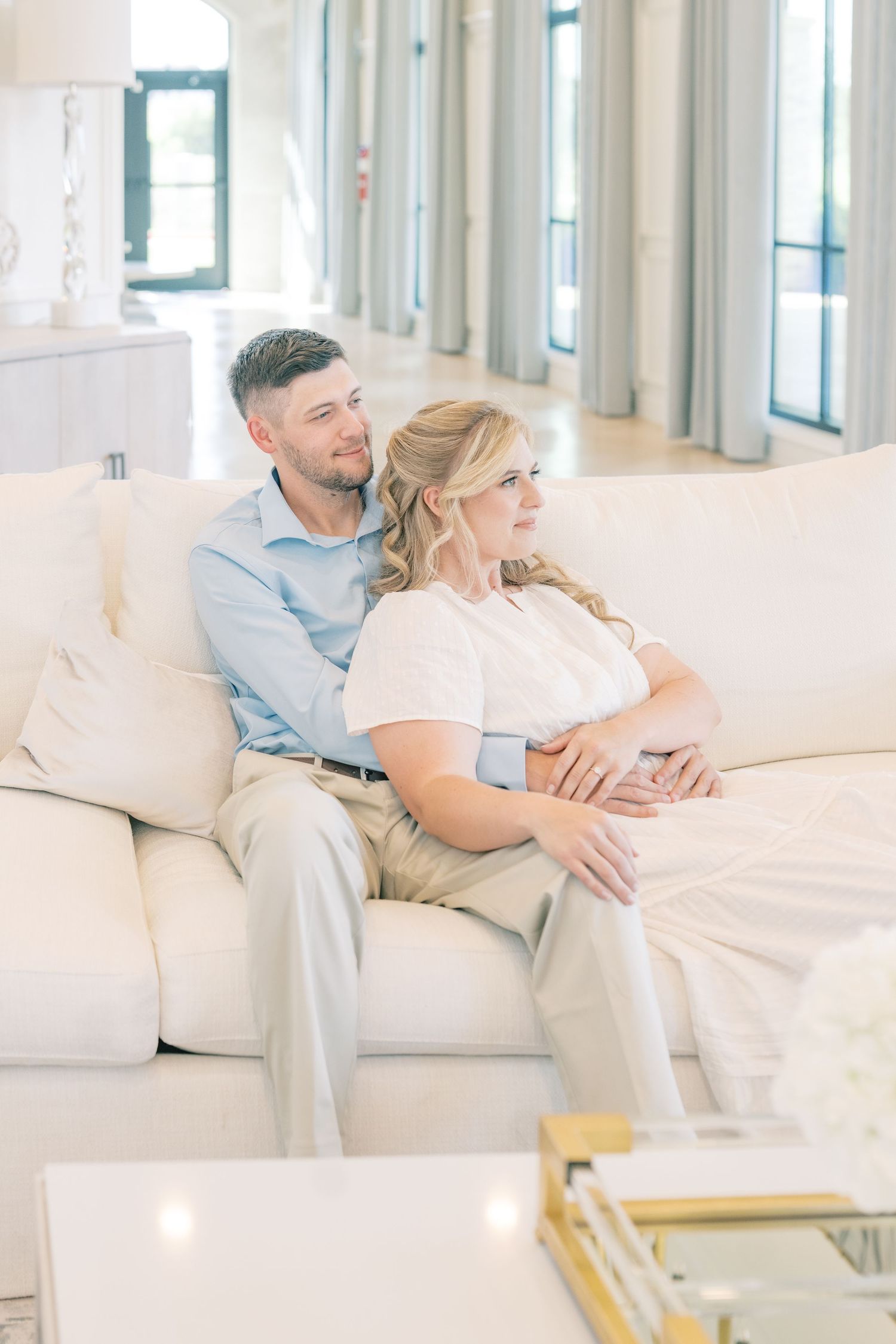 Couple sitting together on a white sofa in a bright elegant living room, man embracing woman from behind.