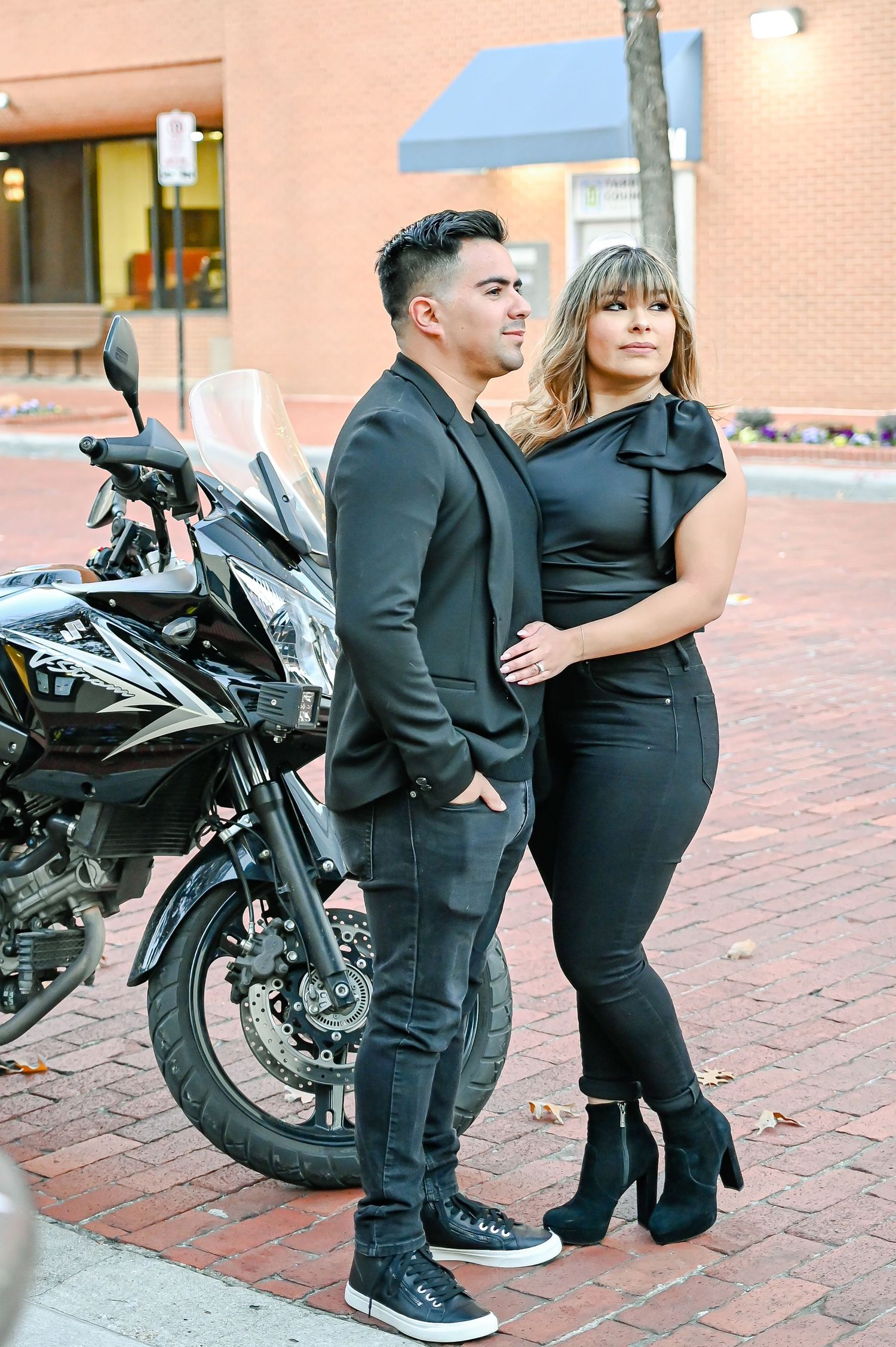 Couple in matching black outfits posing beside a motorcycle on a brick sidewalk.