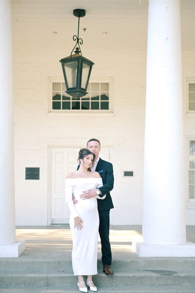 Couple posing at white colonial building entrance, woman in off-shoulder white gown, man in dark suit embracing her.