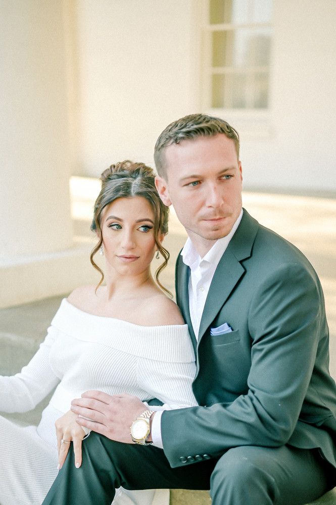 Couple in formal wedding attire sitting closely together outside a sunlit white building.