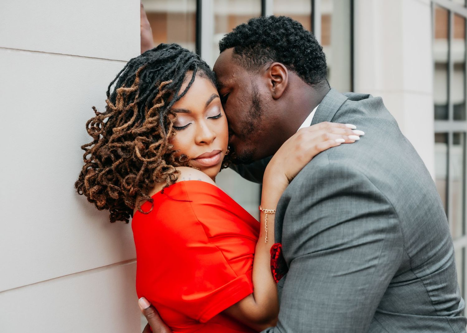 A man in a gray suit kisses a woman in a red dress as she leans against a white wall.
