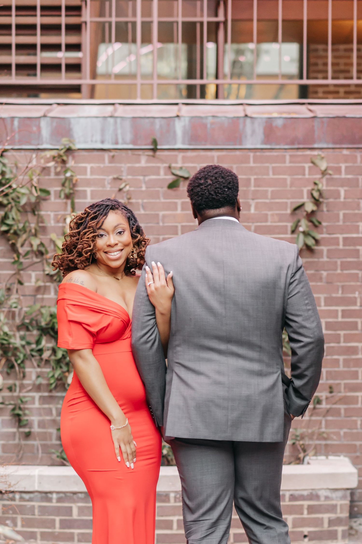 Smiling woman in red dress showing engagement ring beside man in gray suit against brick wall.