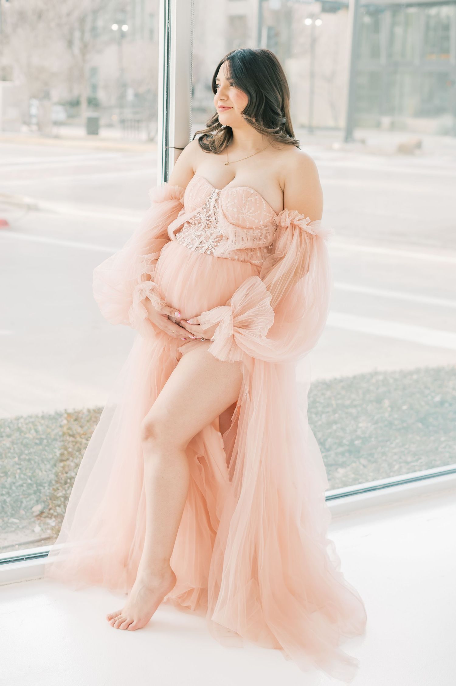 Pregnant woman in a blush pink tulle maternity gown posing by a large window during a maternity photo session.