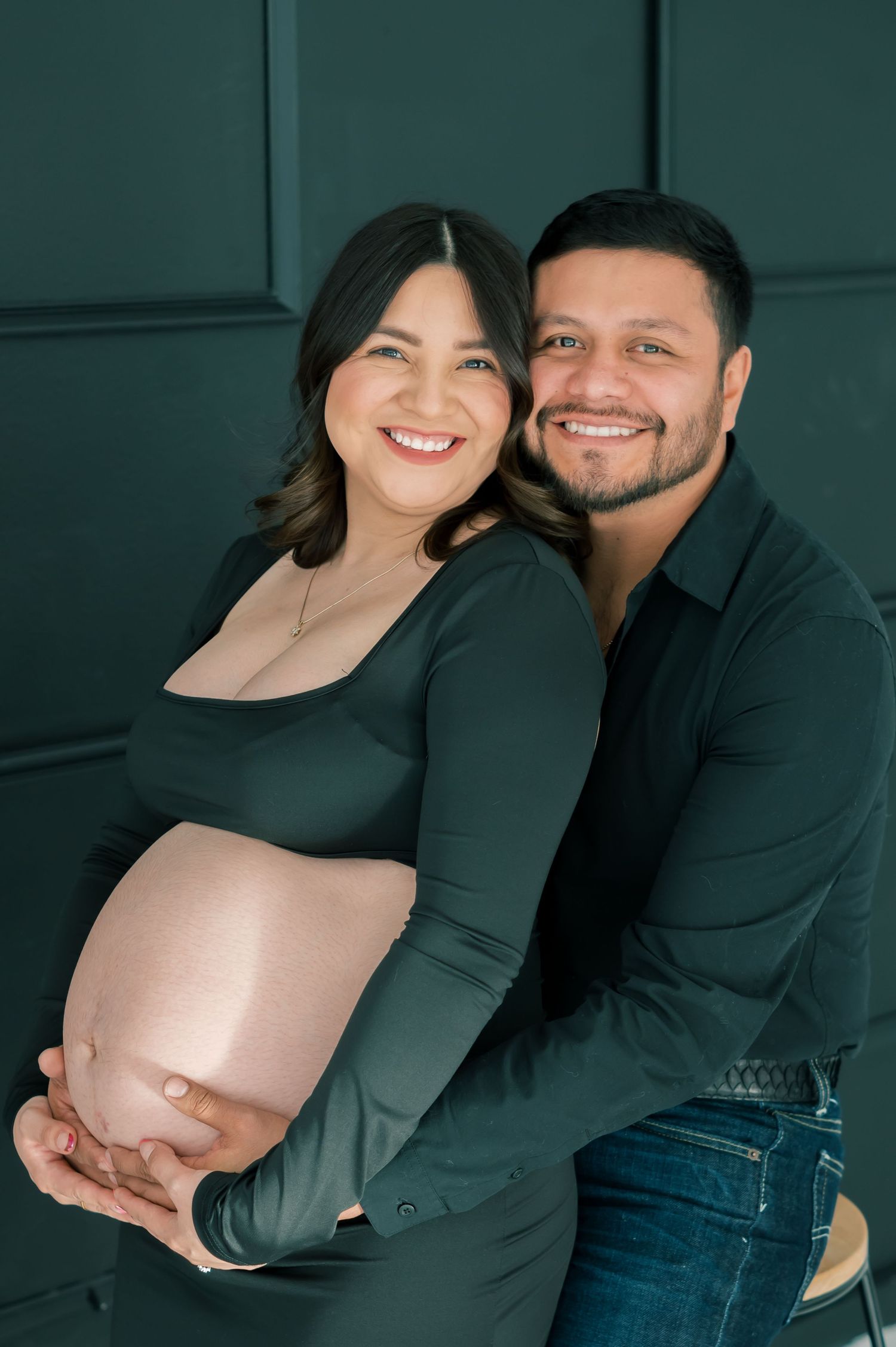 Smiling pregnant couple in matching dark green outfits pose together, man embracing woman from behind as she holds her baby bump.